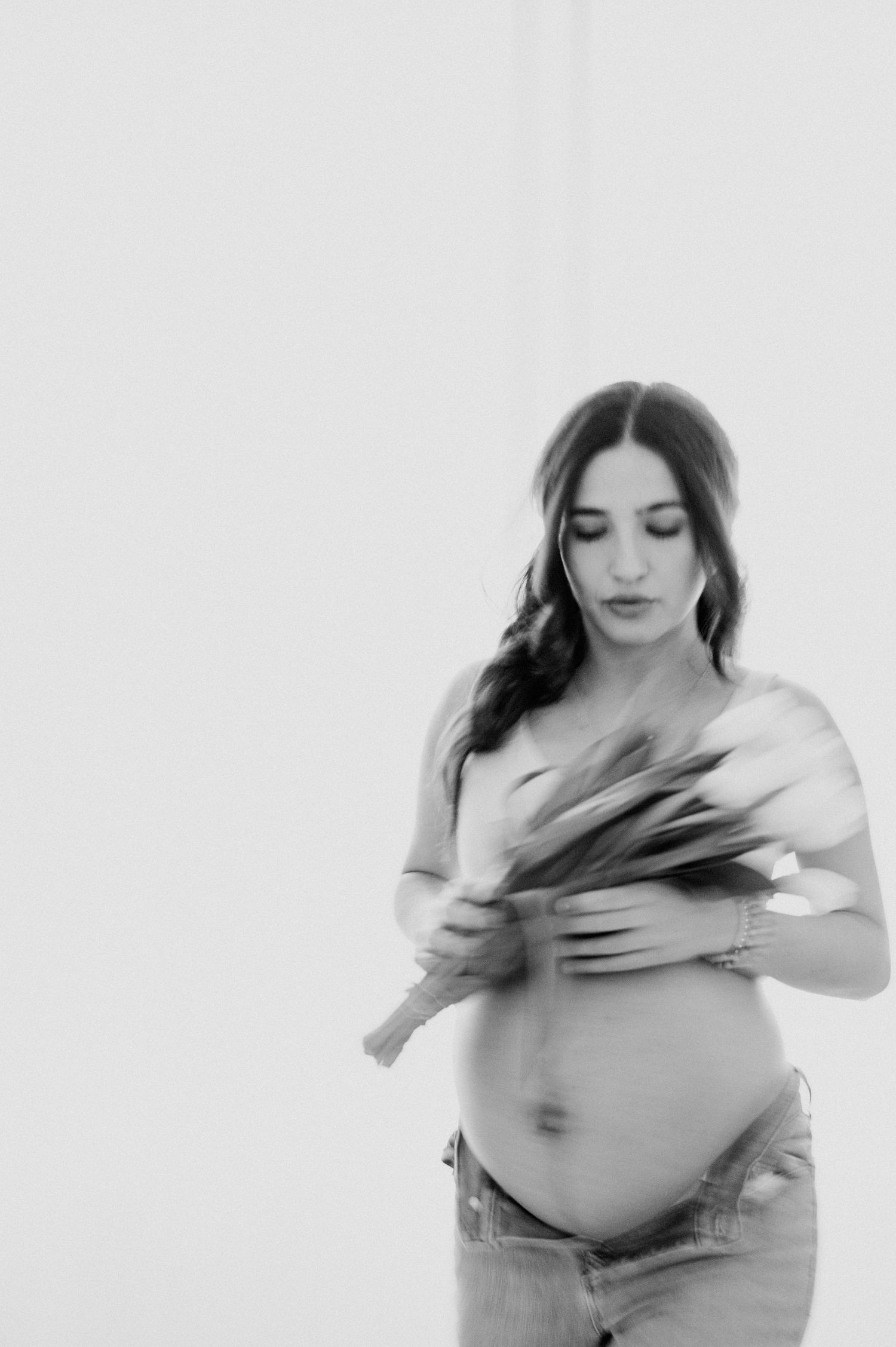Black and white maternity photo of pregnant woman holding flowers, wearing jeans, looking down at baby bump.