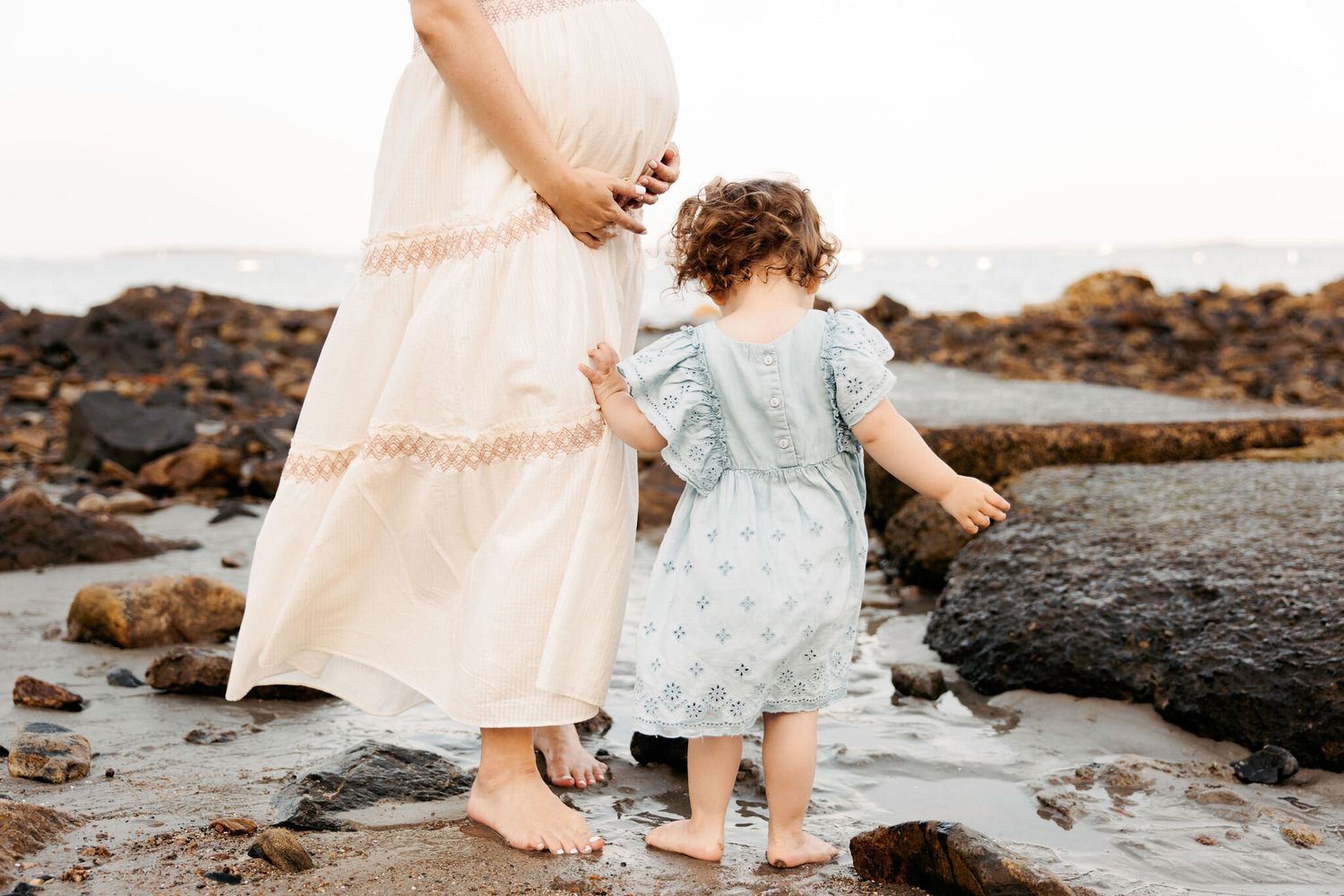 Mother and child walking along rocky shoreline in matching light dresses.