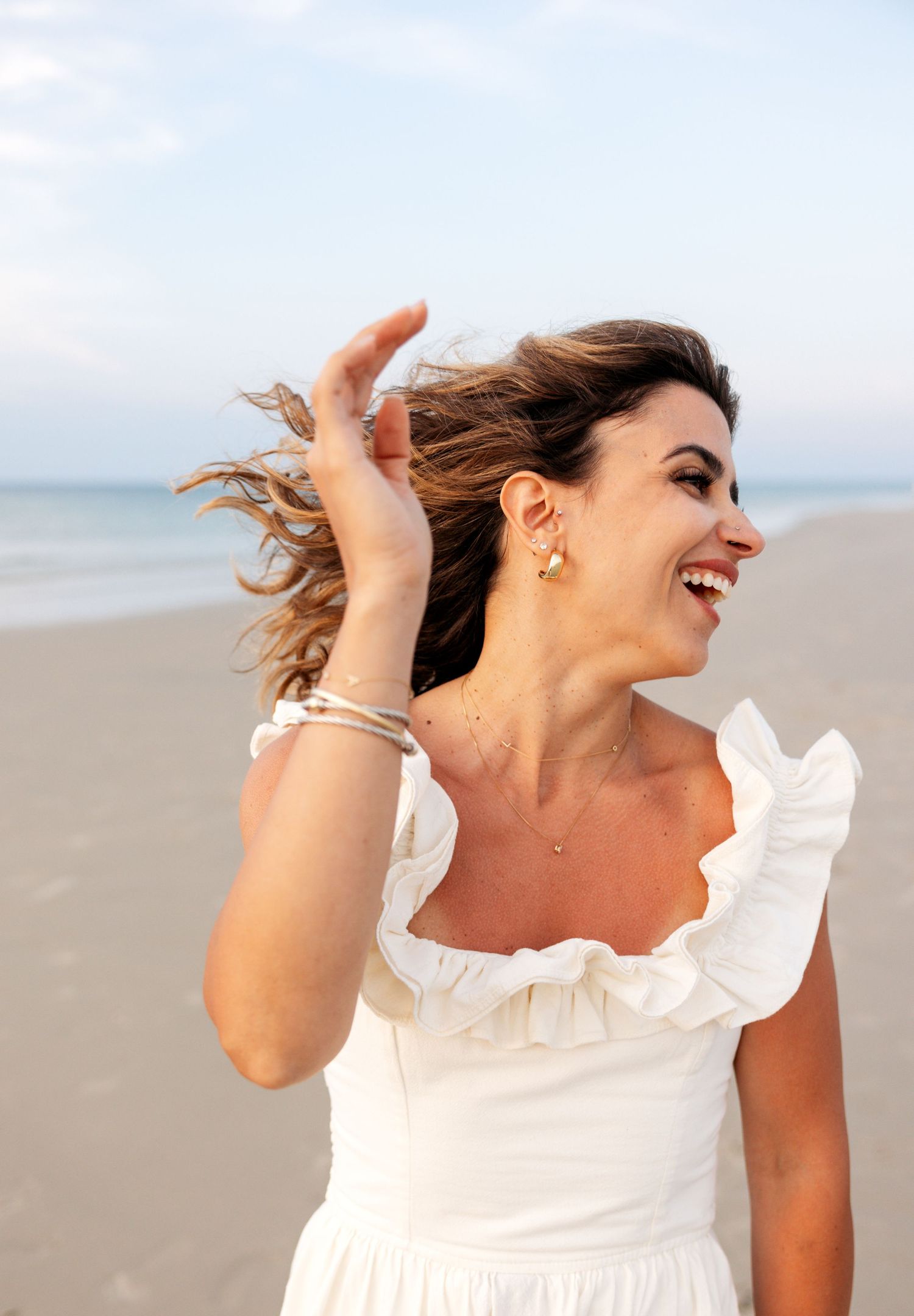 A woman in a white ruffled dress laughs joyfully while walking on a beach during sunset.
