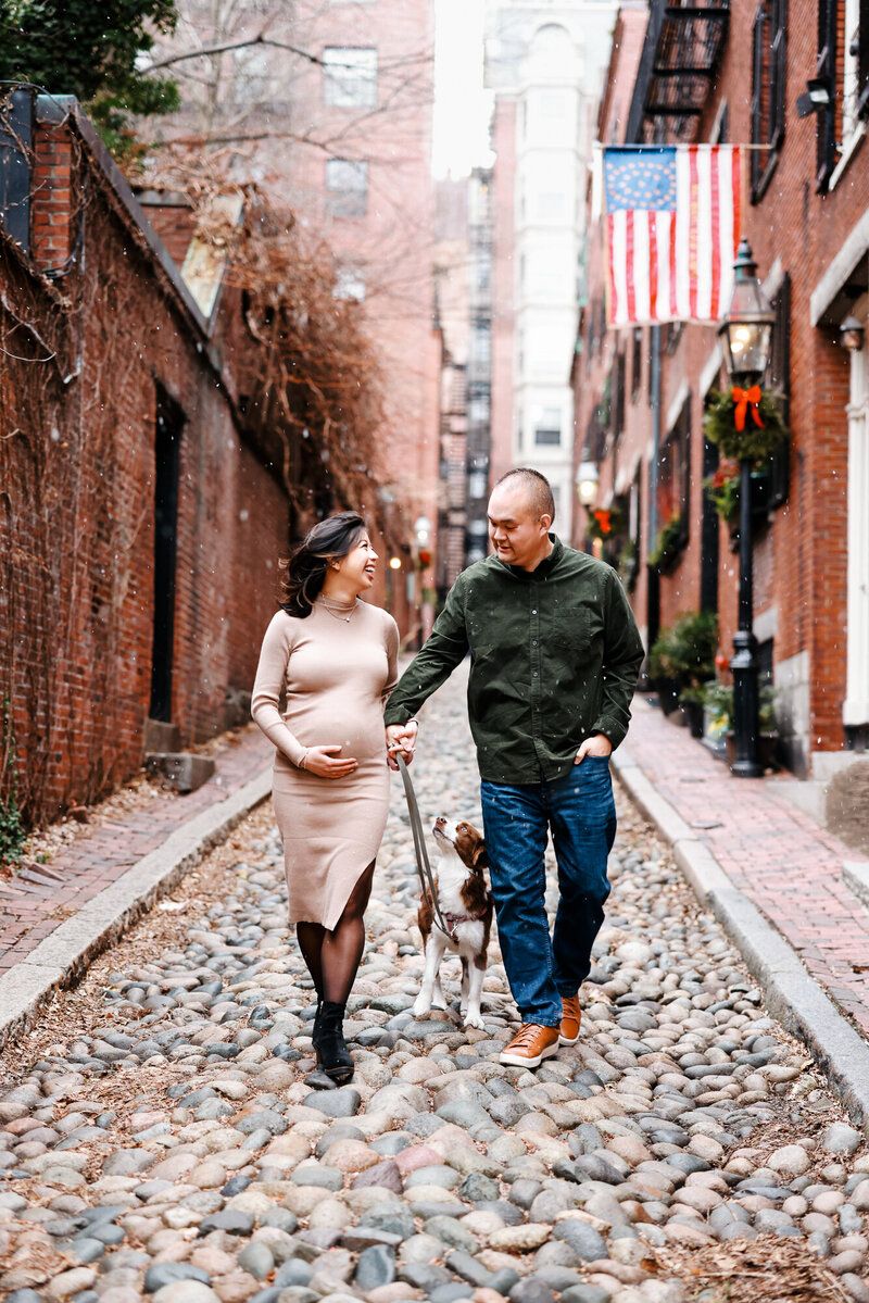Pregnant couple walks dog on historic cobblestone alley in Boston, with brick buildings and American flag backdrop.