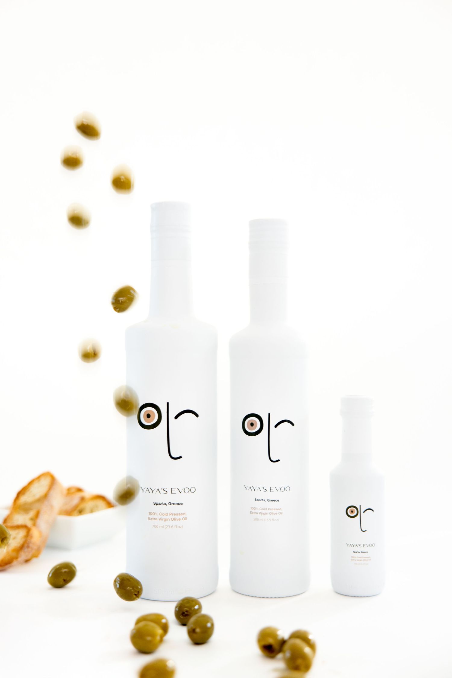 White cosmetic product bottles surrounded by scattered golden olives and olive oil drops on a clean white background.