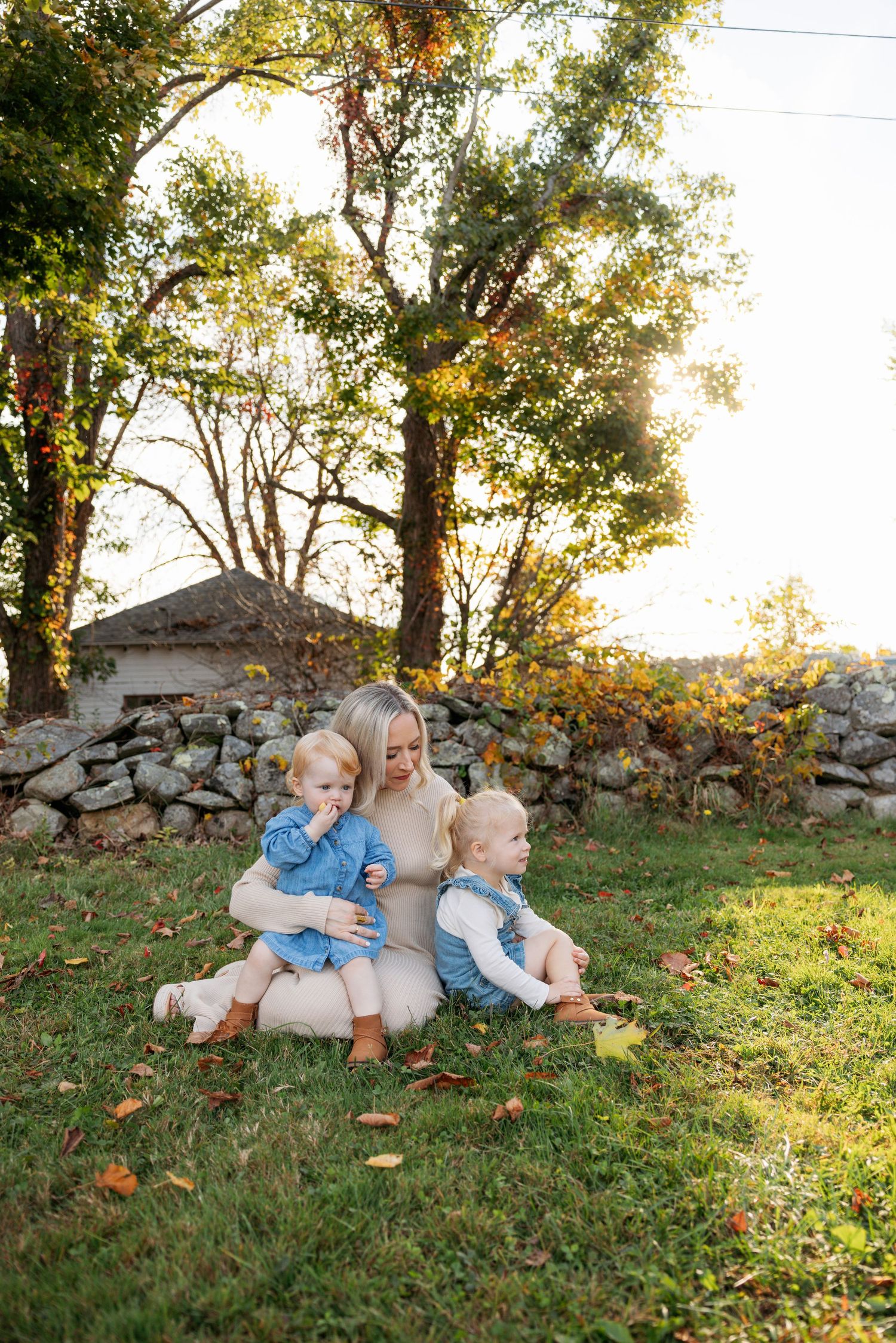 Candid outdoor family moments captured on grass near a stone wall during golden hour with autumn trees in the background.