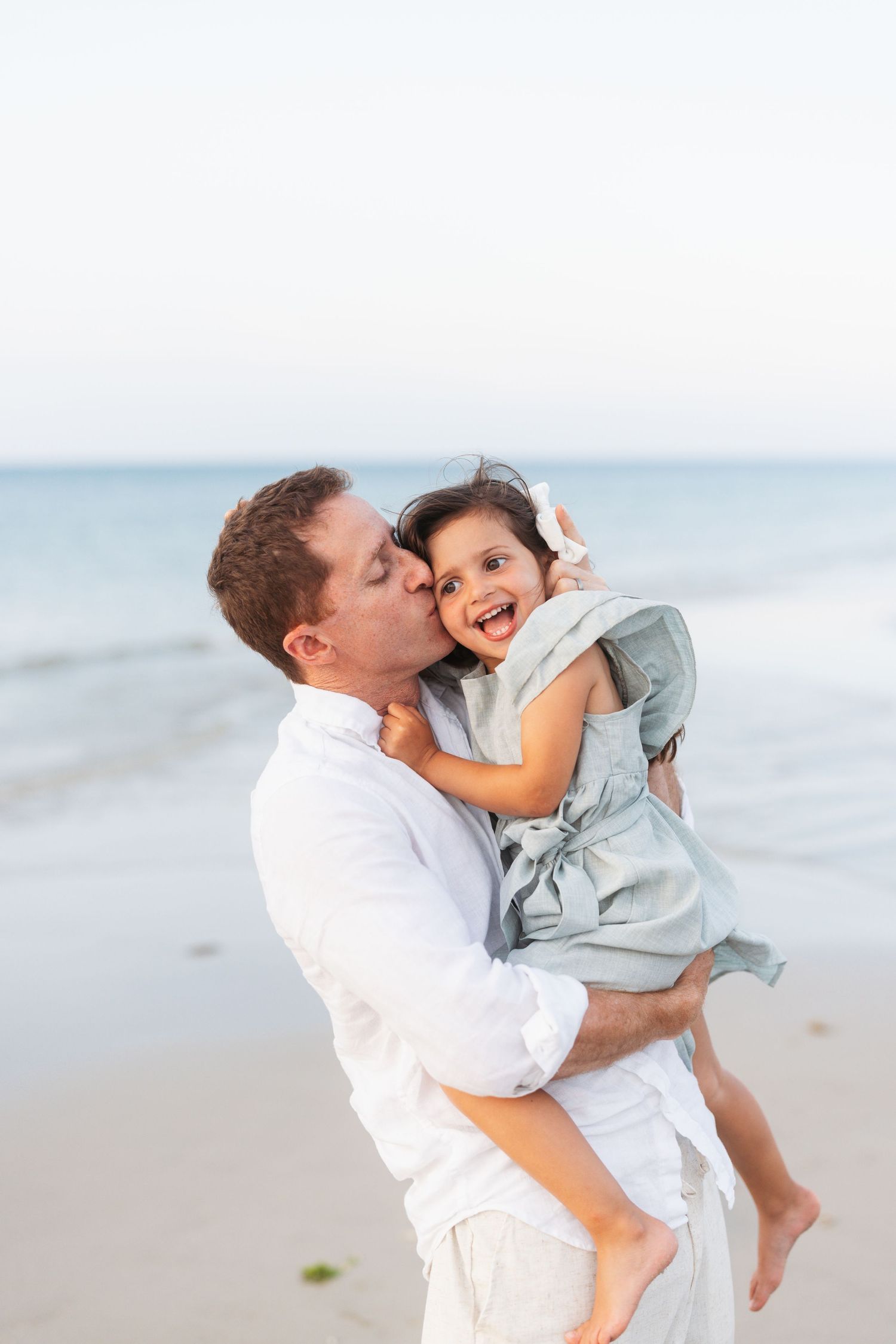 A series of connected photos shows playful moments between parent and child at the beach with matching white outfits.