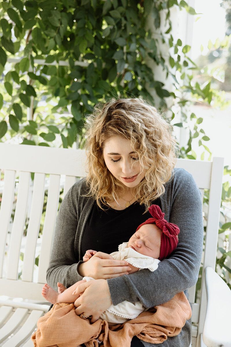Tender moment on white porch bench with baby under leafy green foliage.