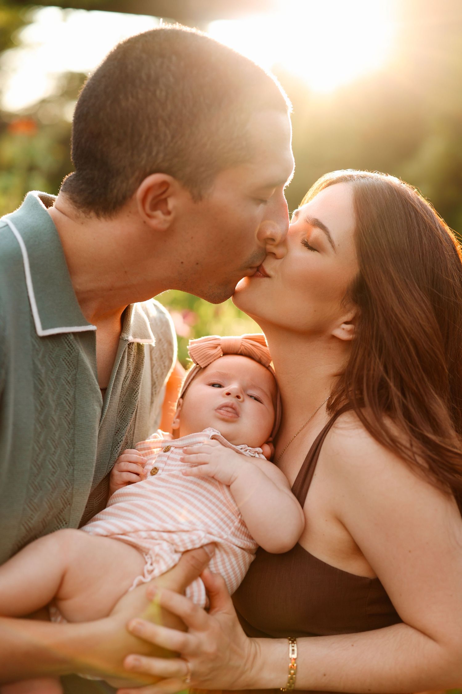 Parents kissing while holding their newborn baby outdoors in warm golden sunset light.