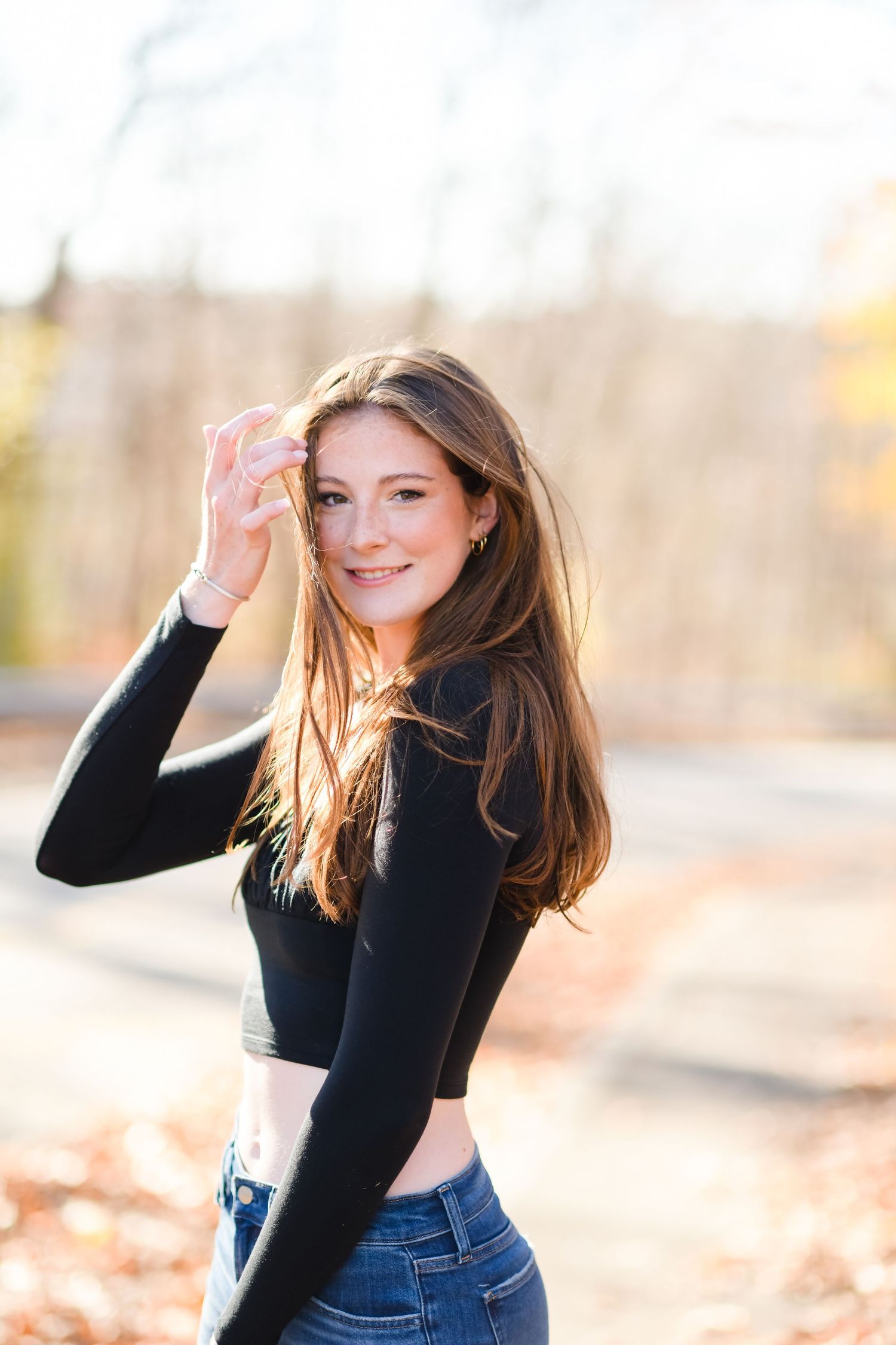 A person in a black long-sleeve top poses outdoors during golden hour with autumn foliage in the background.