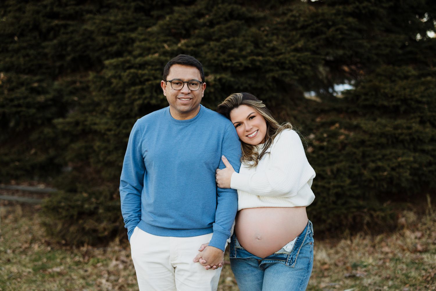 Couple embraces during outdoor maternity photoshoot with evergreen trees in background.