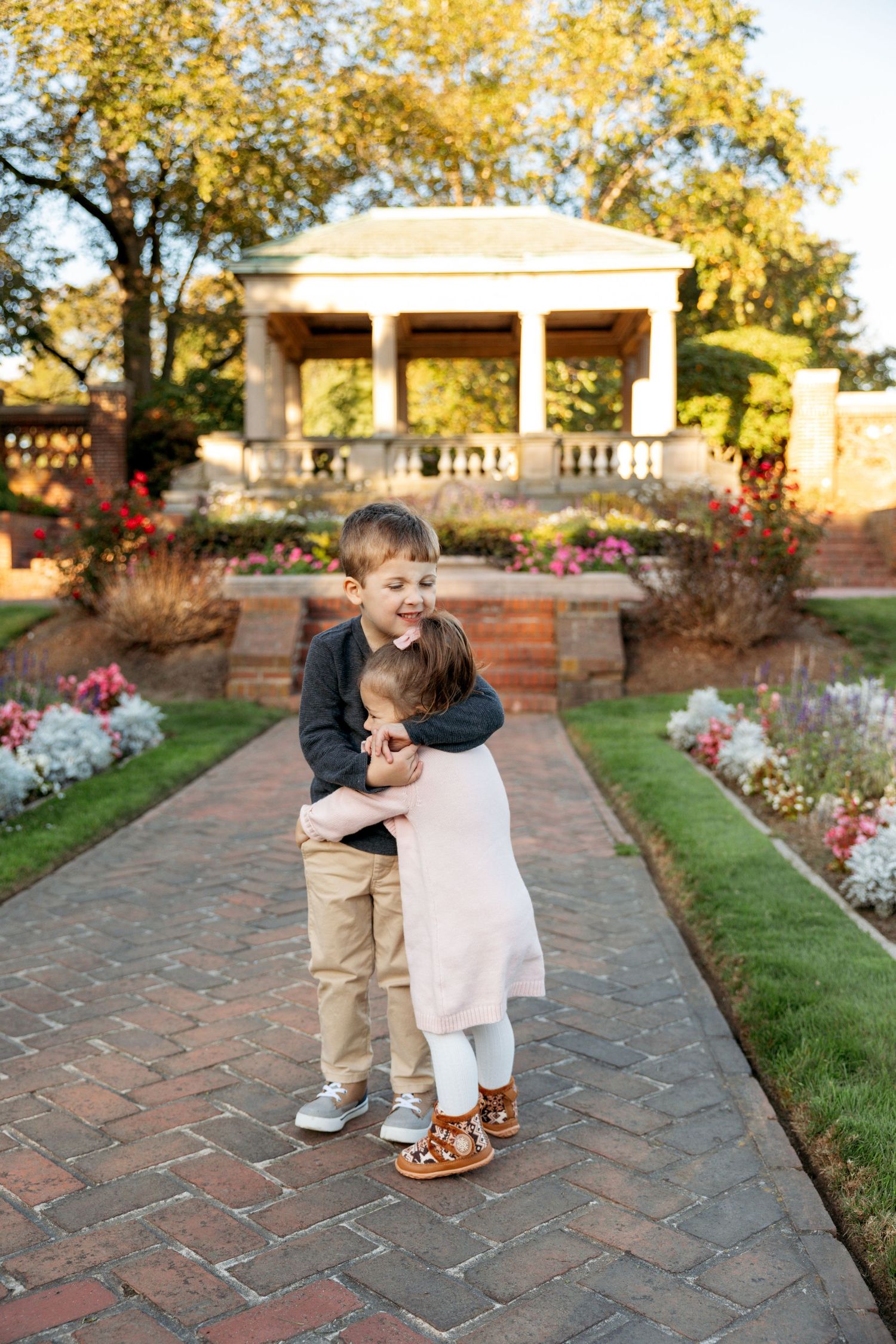 Two children share a sweet moment walking together on a brick pathway surrounded by flowers at sunset.