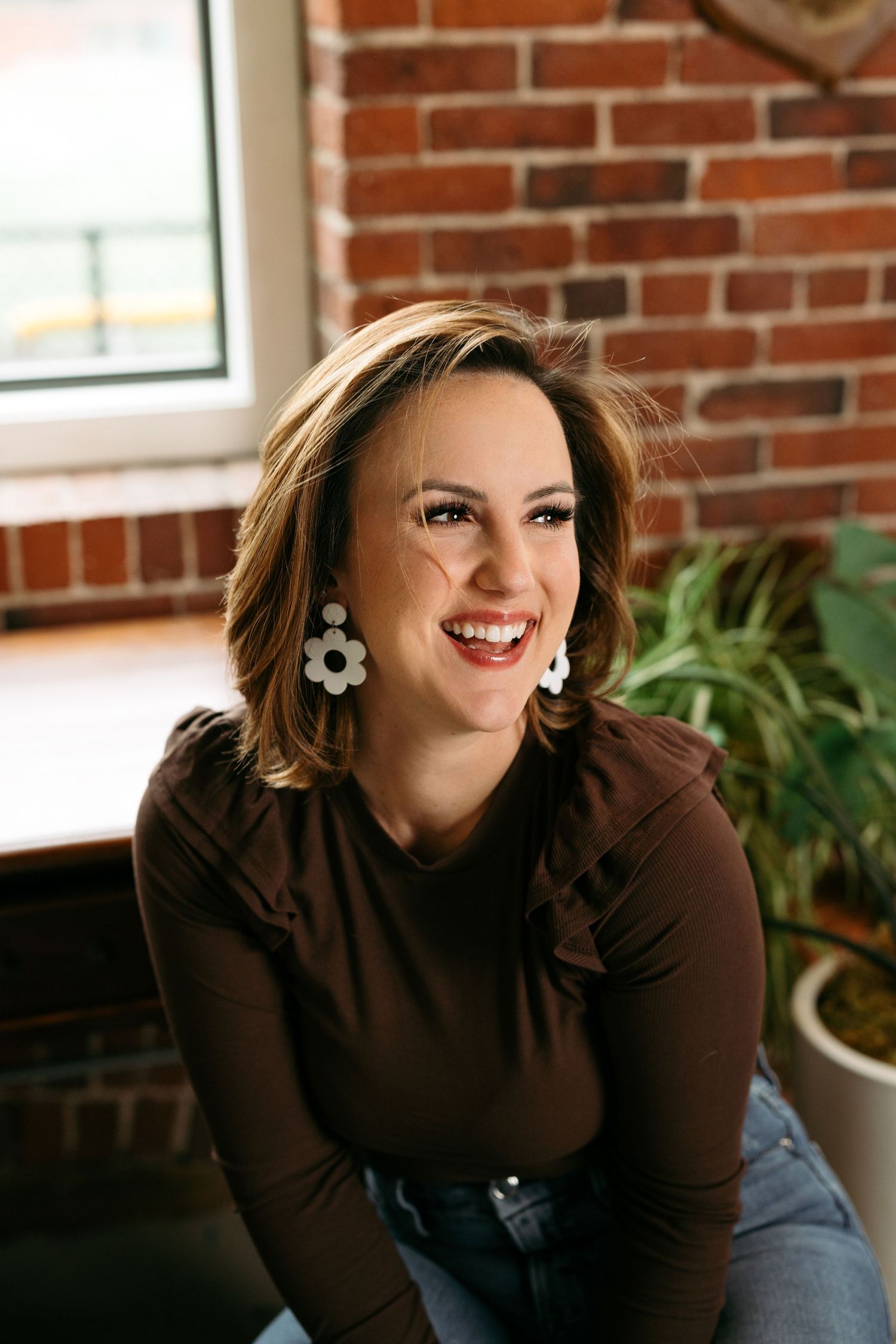 Smiling woman with short brown hair wearing white flower earrings and brown top, sitting near brick wall and plants.