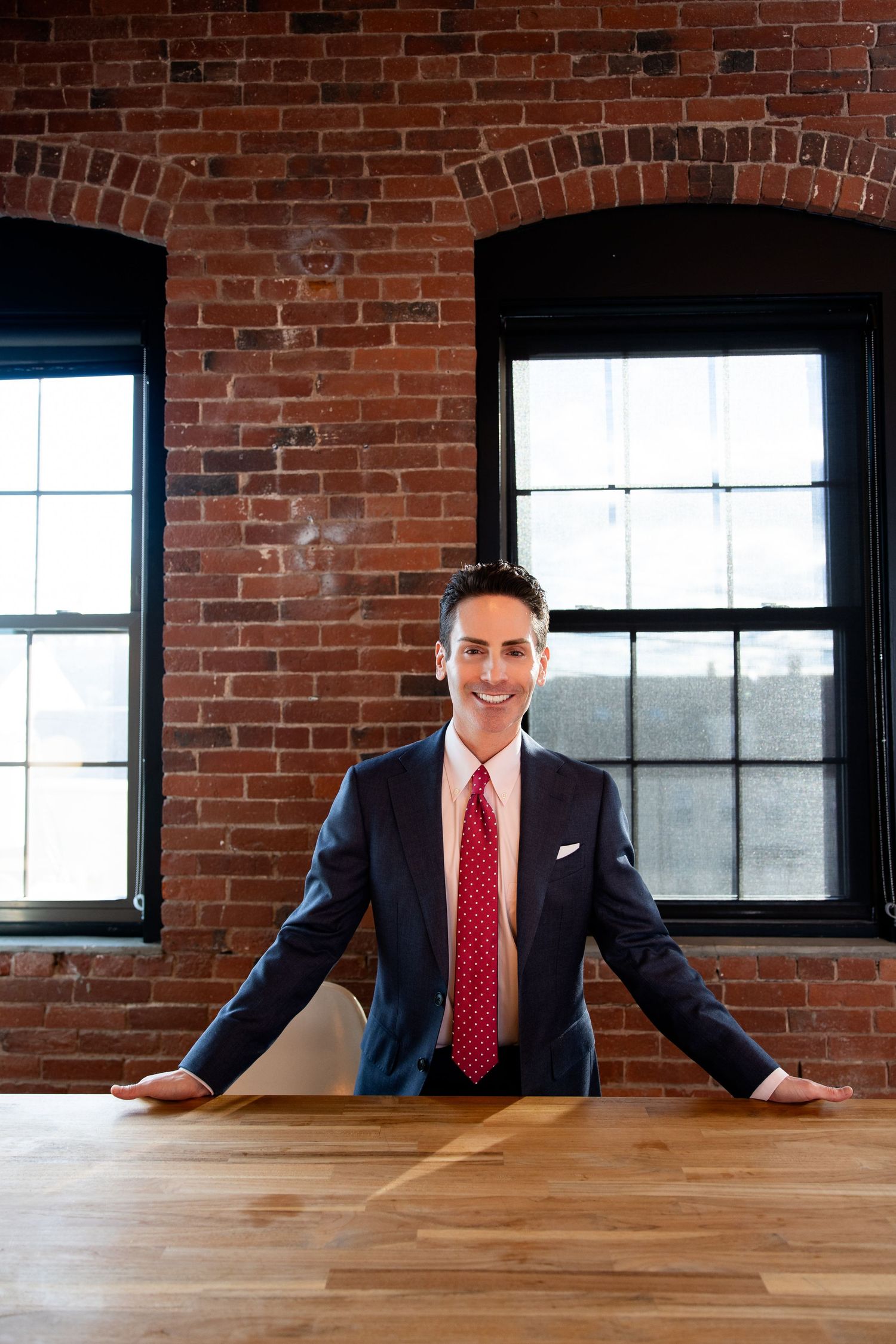 Smiling businessman in navy suit and red tie stands confidently at wooden table in brick-walled office.