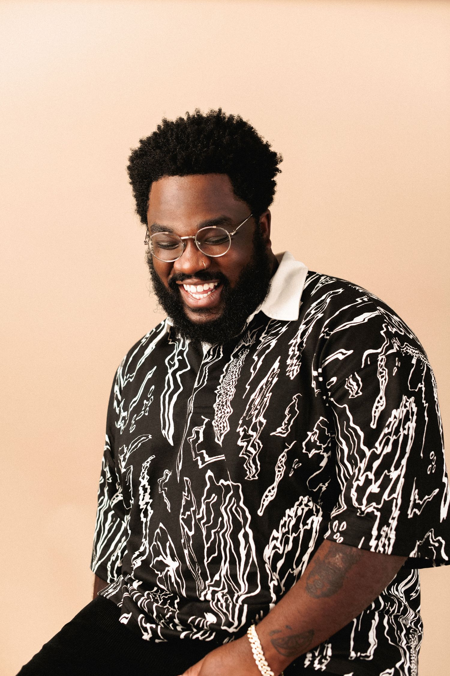 Smiling bearded Black man wearing glasses and patterned shirt against beige background.