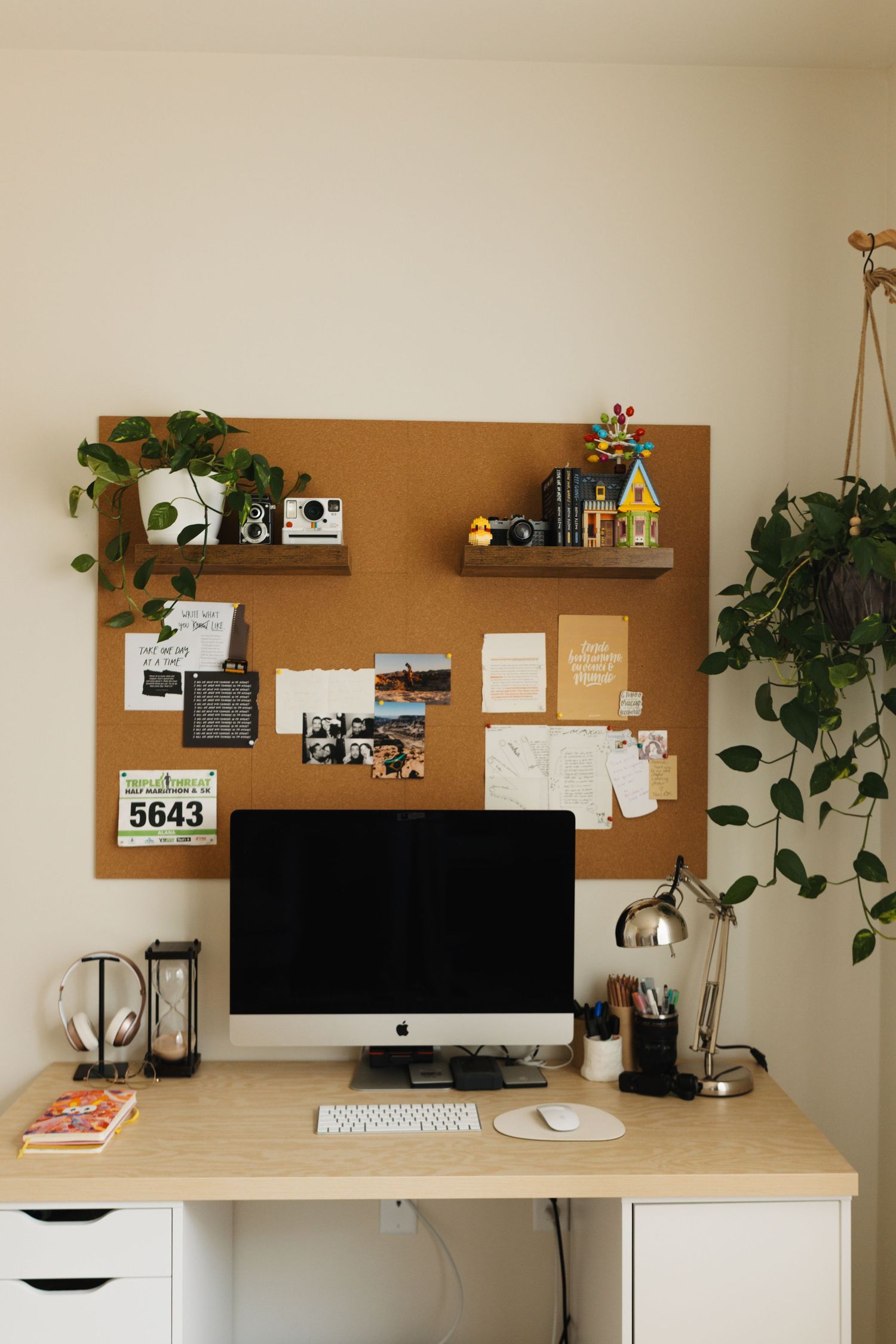 A minimalist home office workspace with cork board, wooden desk, iMac computer, and hanging plants against beige walls.