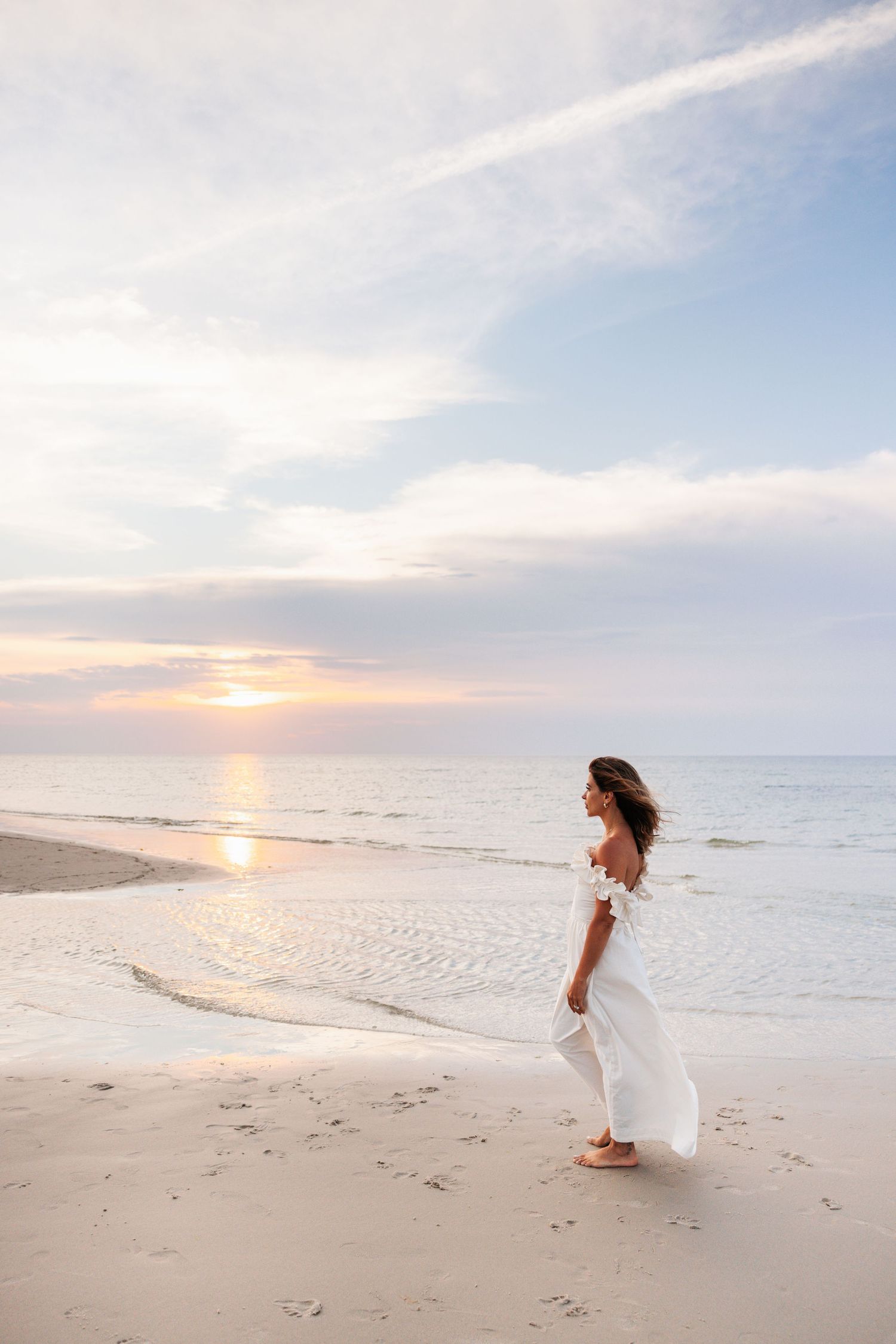 A figure in a flowing white dress walks along an empty beach at sunset with waves lapping the shore.