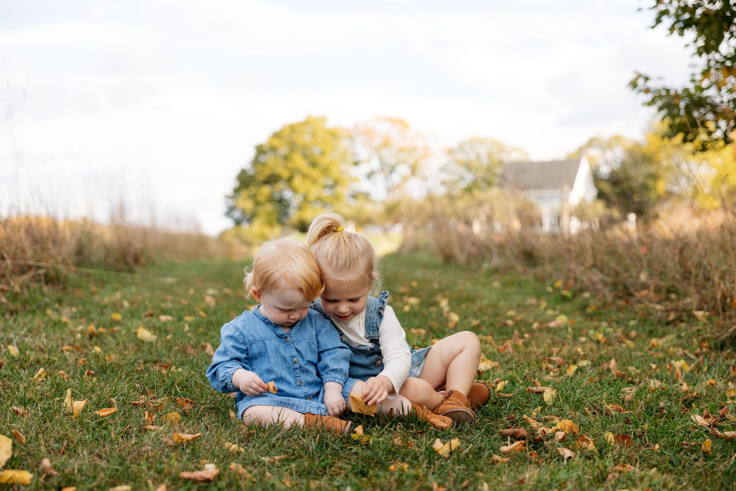 A couple sits together on grass scattered with fallen autumn leaves during a photoshoot.