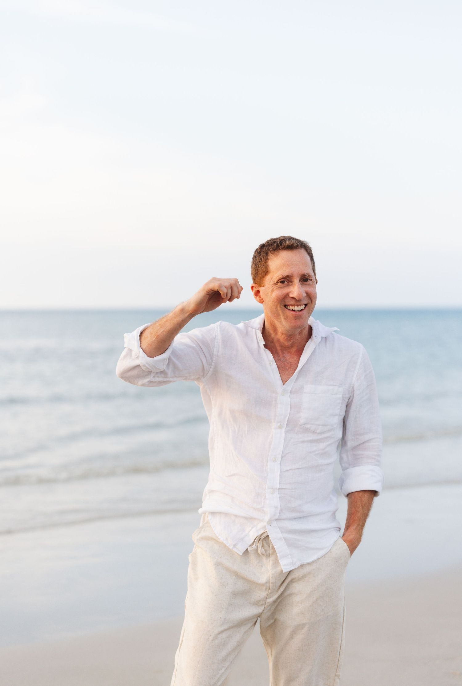 Person in white shirt and light pants dances joyfully along a sunny beach with ocean waves in background.