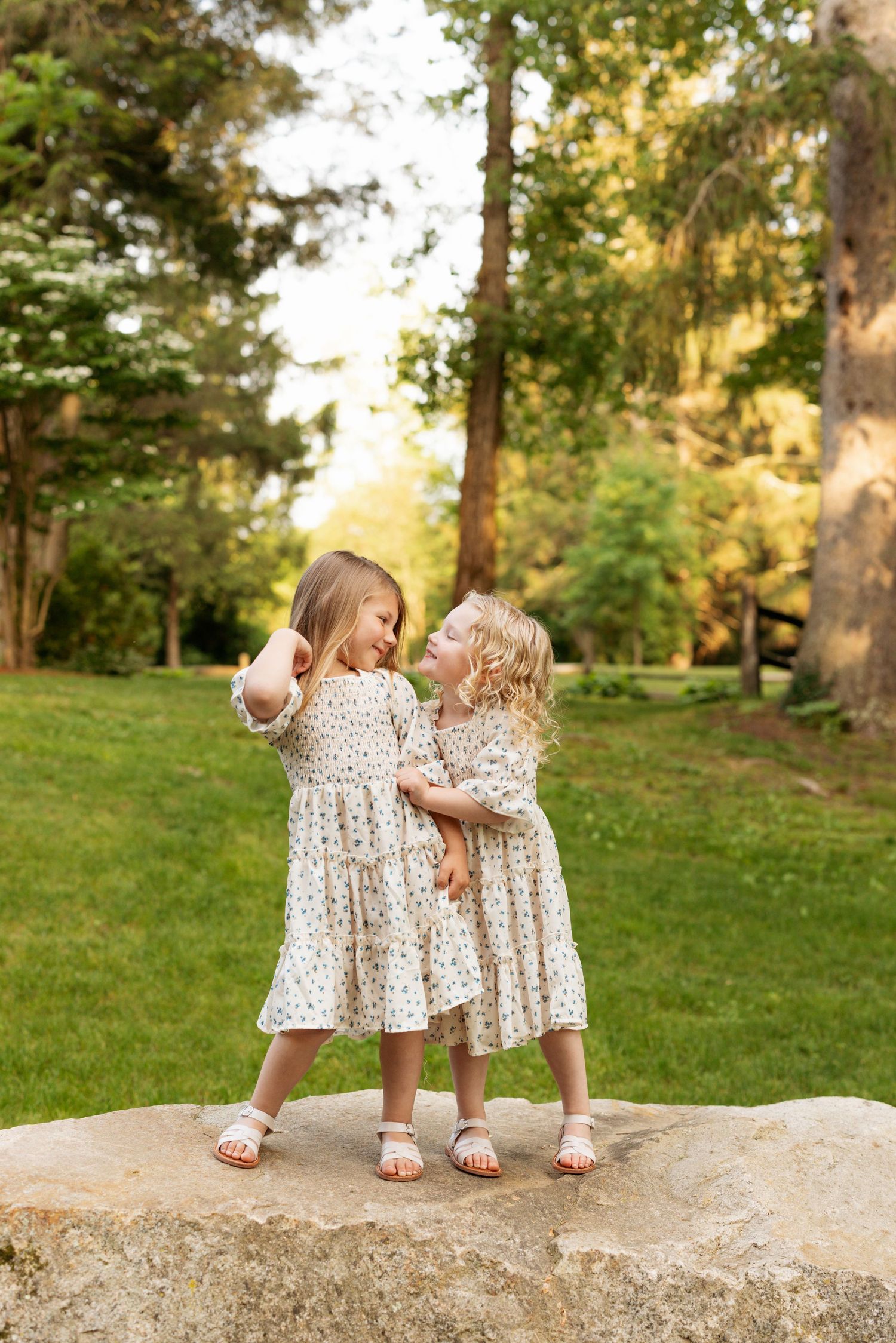 Three small children in matching floral dresses playing on a rock in a lush park setting.