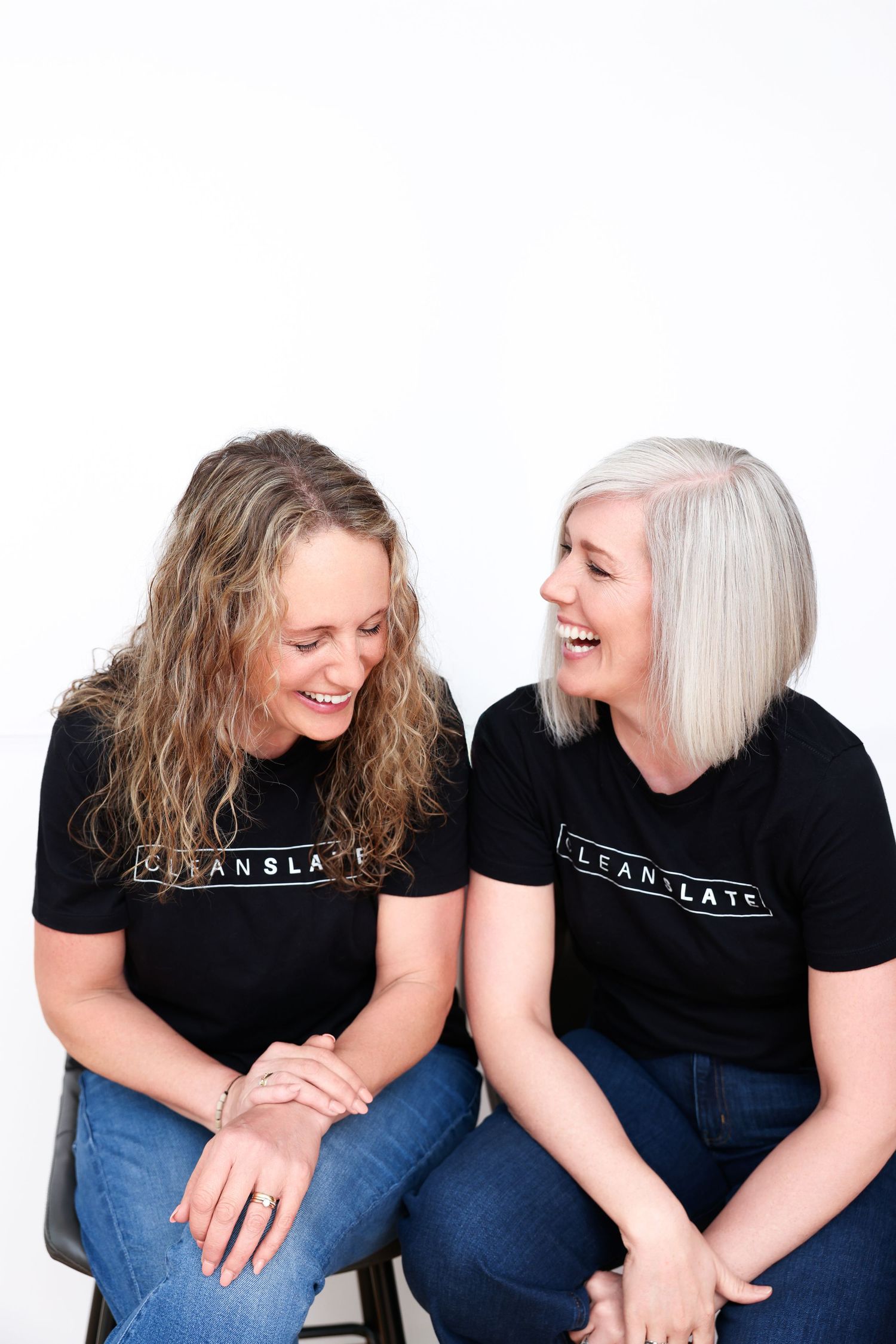 Two women wearing matching black Clean Slate t-shirts sit together laughing and smiling against a white background.