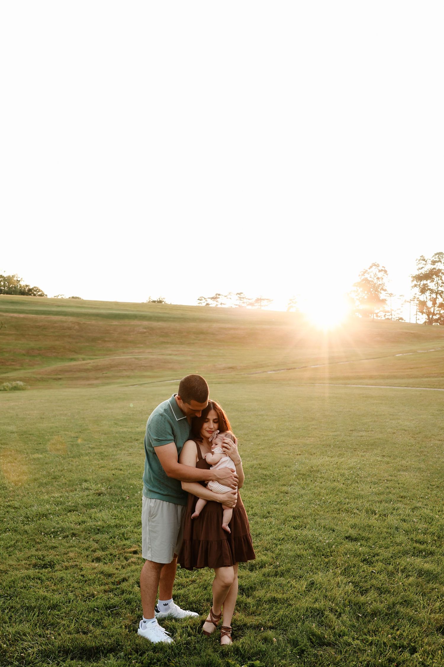 Couple holding newborn baby in a sunlit green field at golden hour sunset.