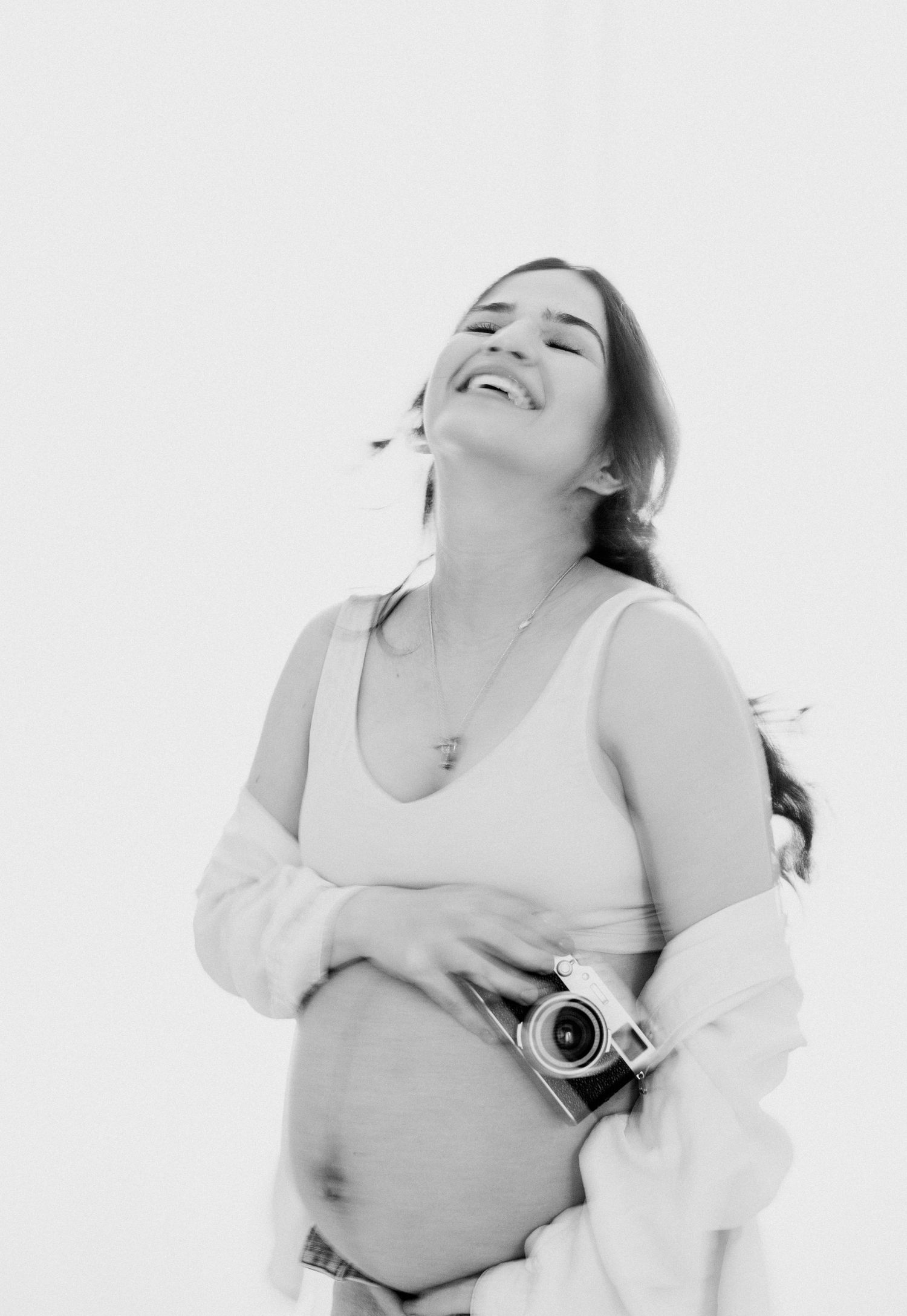 Black and white photo of a smiling pregnant woman holding a vintage camera, wearing a tank top and open shirt.