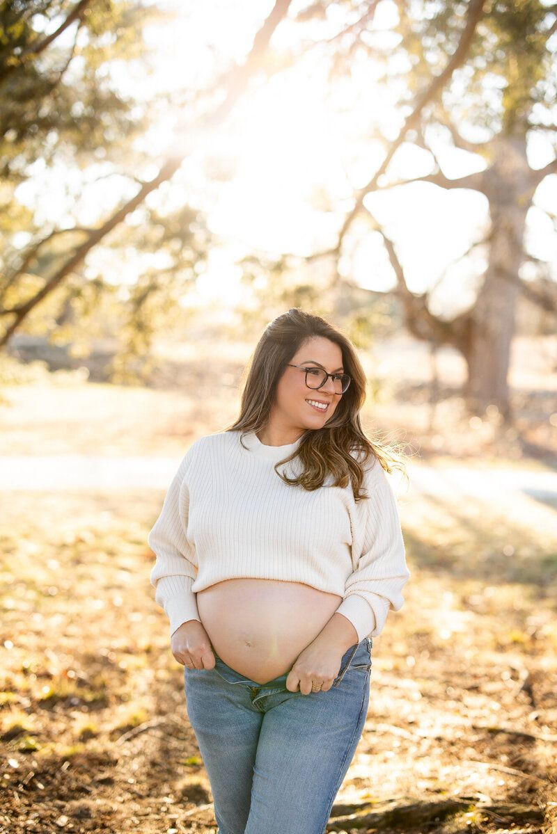 Person in white sweater and jeans poses in autumn sunlight showing pregnant belly.