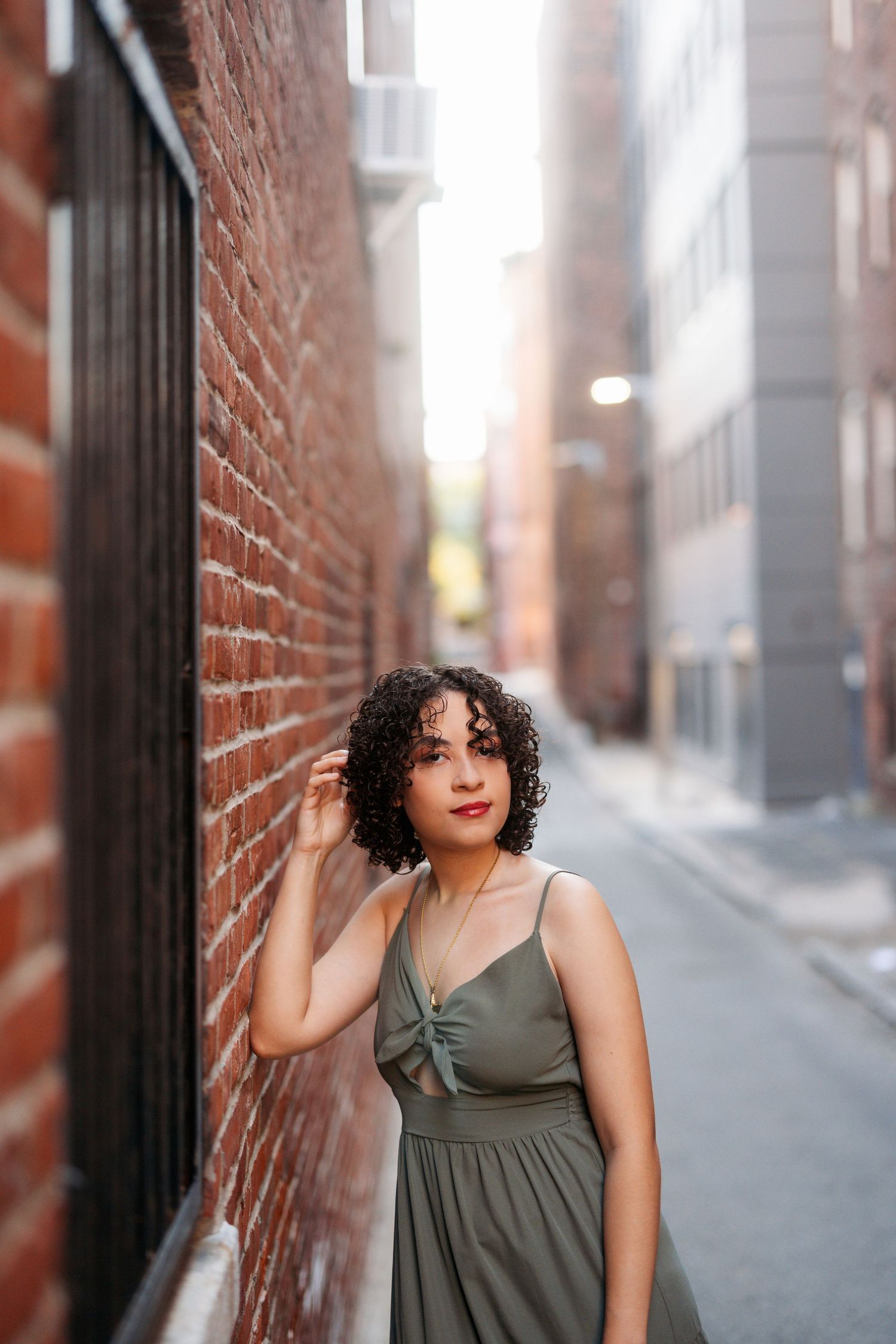 A person in an olive green dress poses against a brick wall in an urban alley with soft natural lighting.