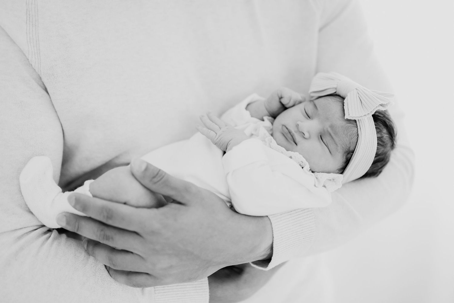 Black and white photo of a sleeping newborn baby cradled in an adults arms, wearing a white bow headband.