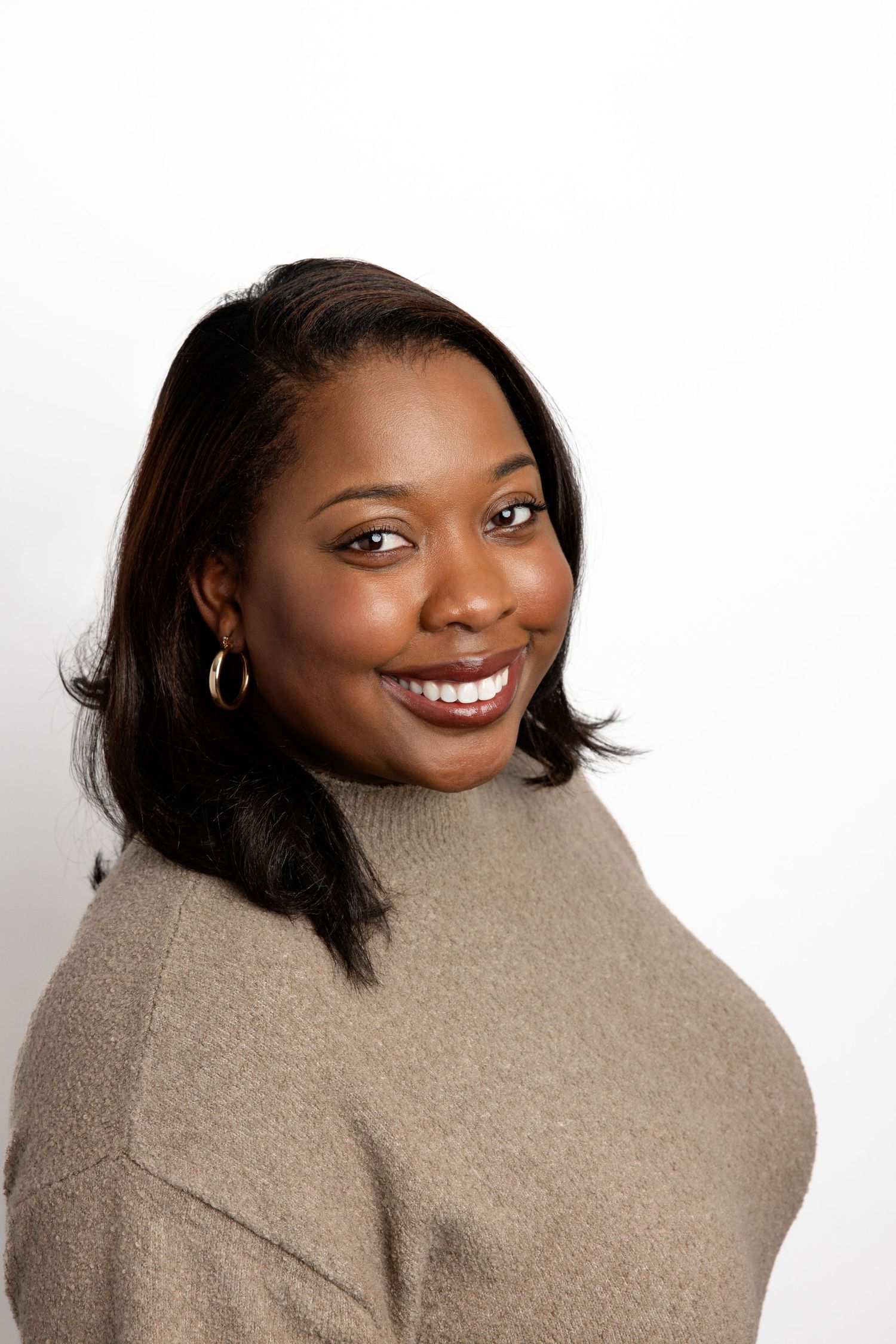 Smiling Black woman with short dark hair wearing a tan turtleneck sweater and gold earrings against a white background.