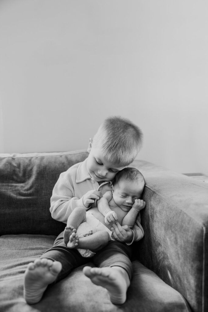 Black and white photo of two young siblings sitting together on a couch, with the older one holding the baby tenderly.