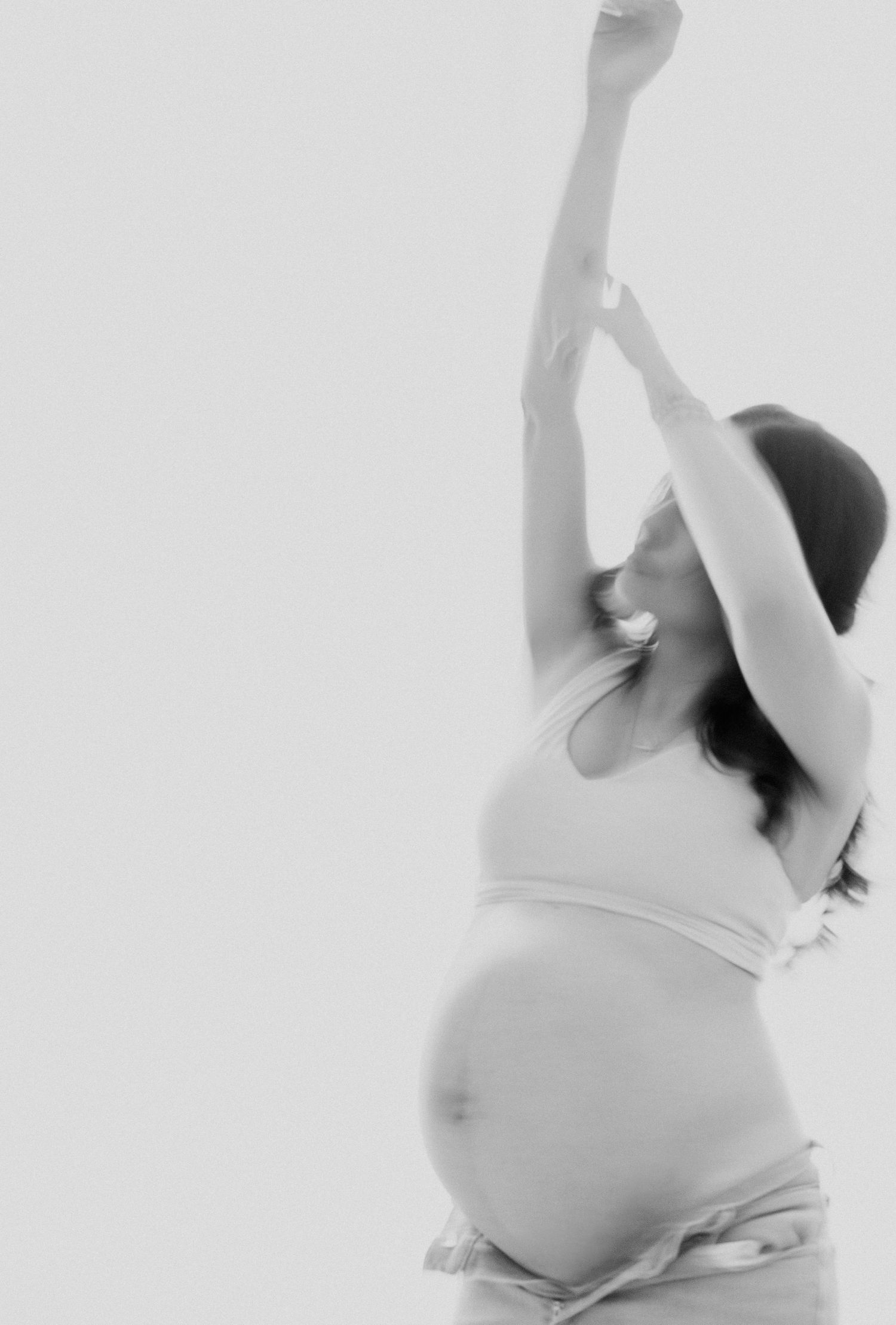 Black and white photo of a pregnant woman in a crop top stretching her arms overhead against a white background.