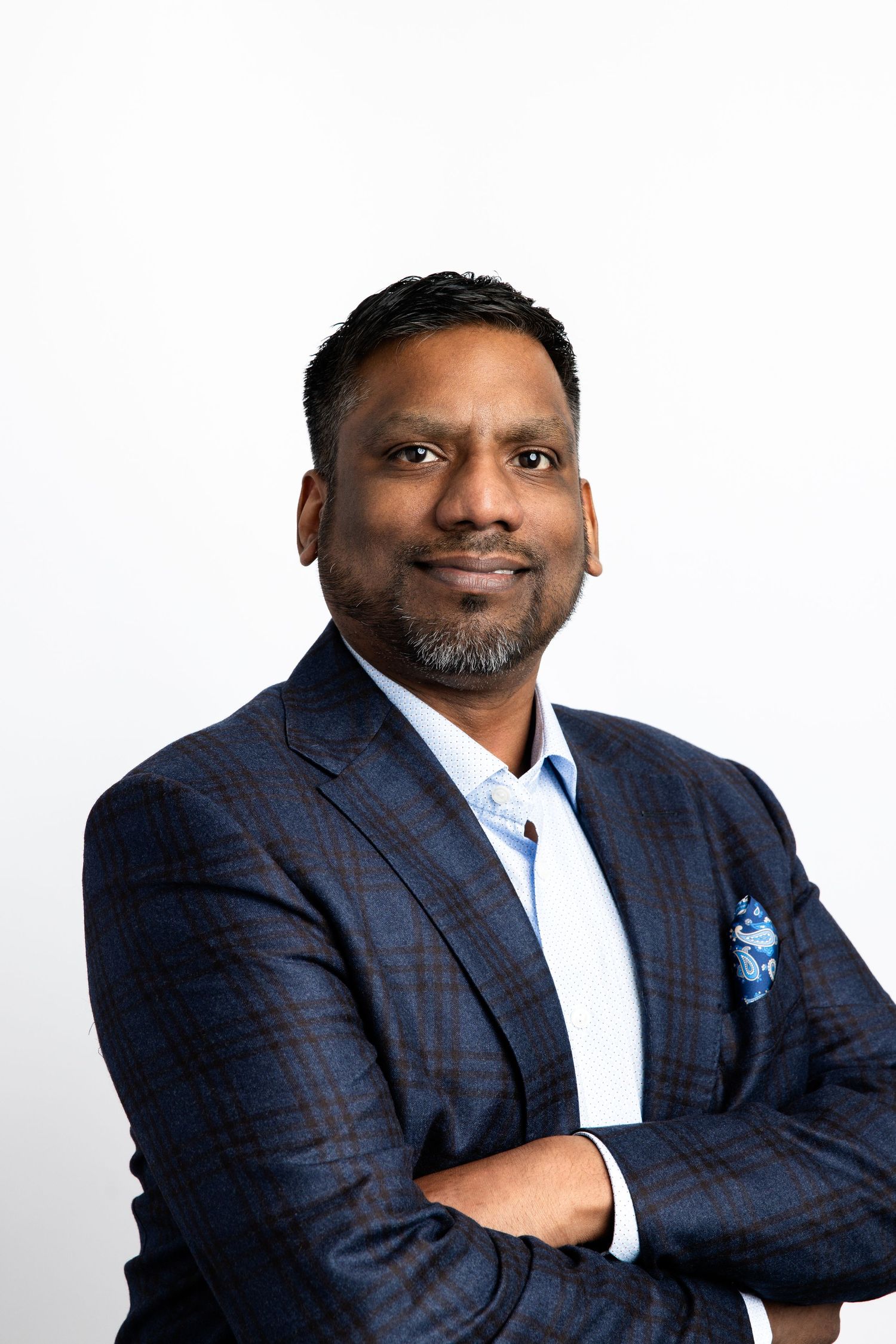 Professional man in navy plaid blazer with blue pocket square, arms crossed, smiling confidently against white background.