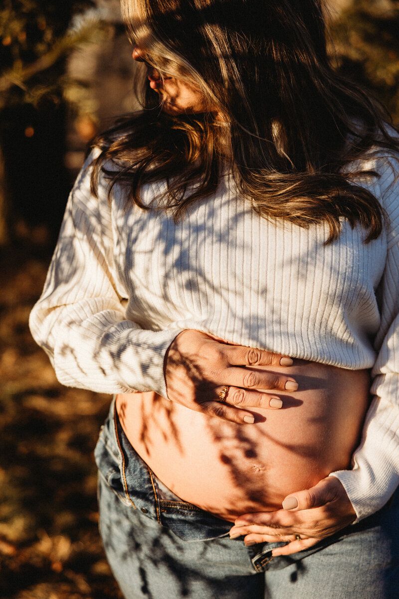 Close-up of hands cradling a pregnant belly in gentle sunlight with dappled shadows.
