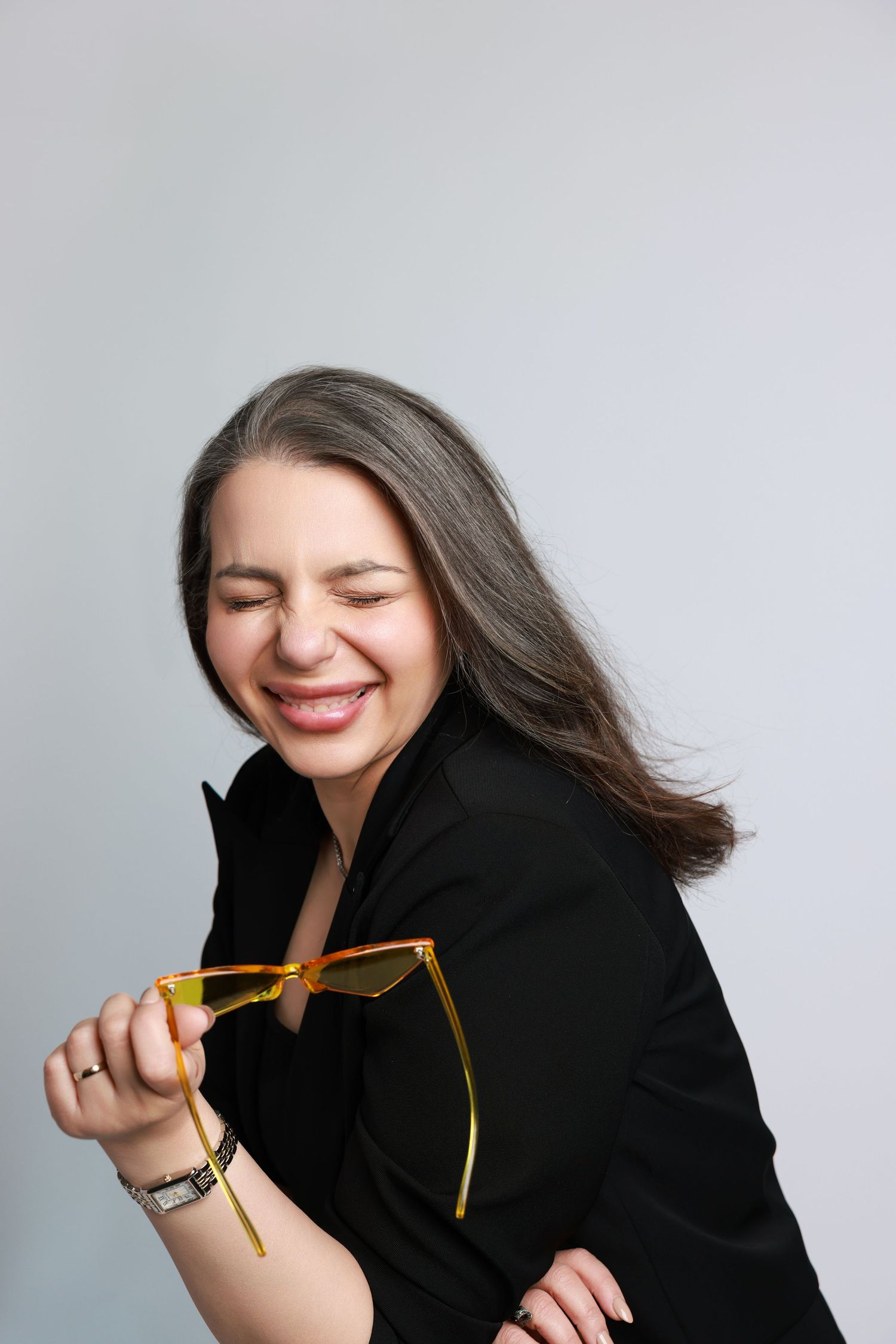A person in a black blazer playfully poses with glasses against a white background.
