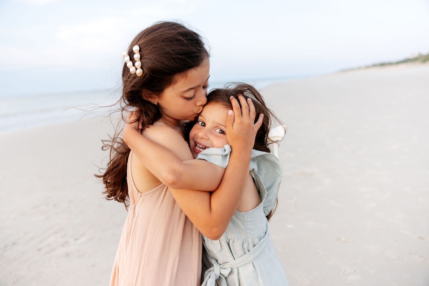 A romantic couple shares tender moments together on a sunny beach in matching light-colored outfits.