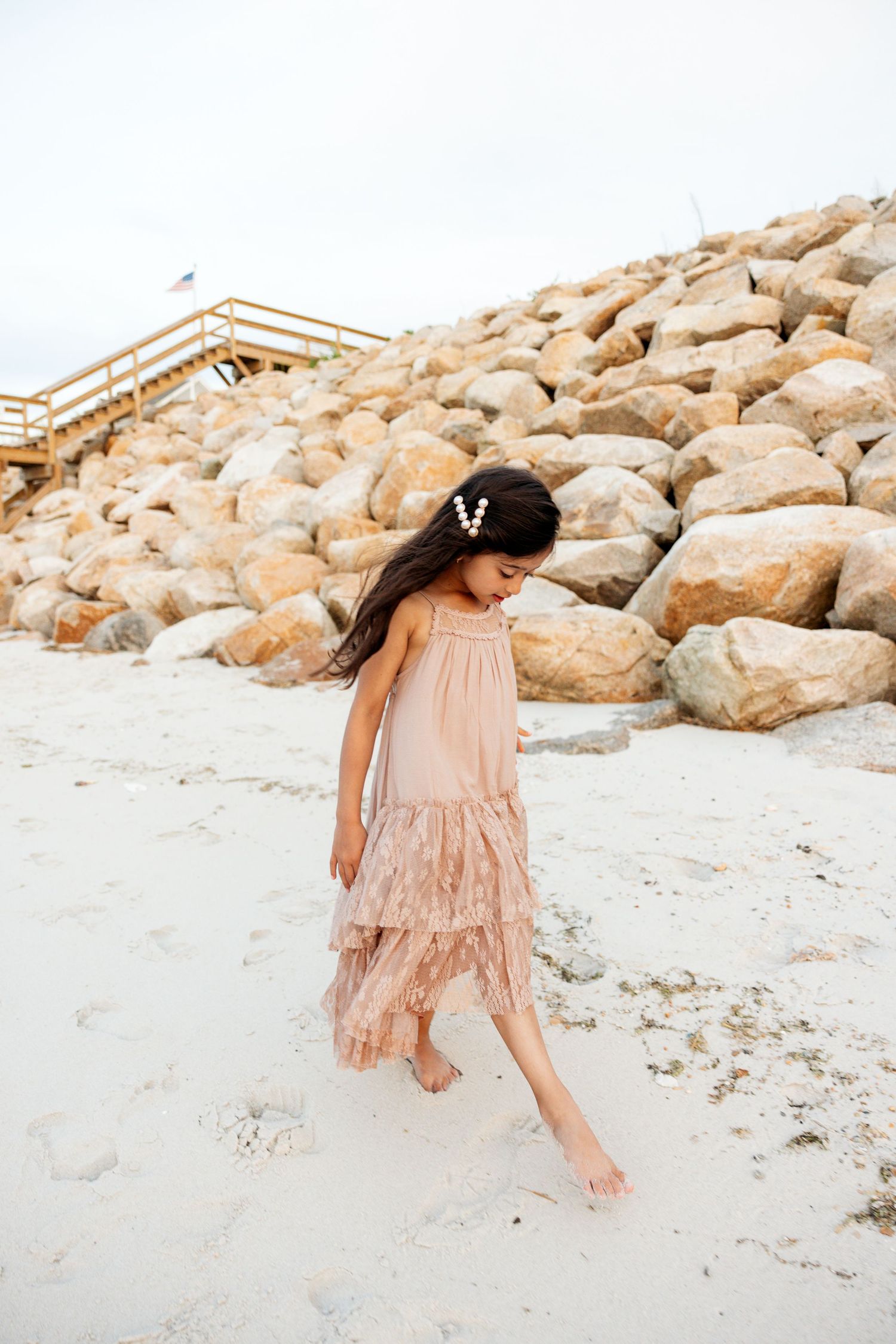 A dreamy beige dress flows gracefully against a sandy beach backdrop with rocky boulders in the background.