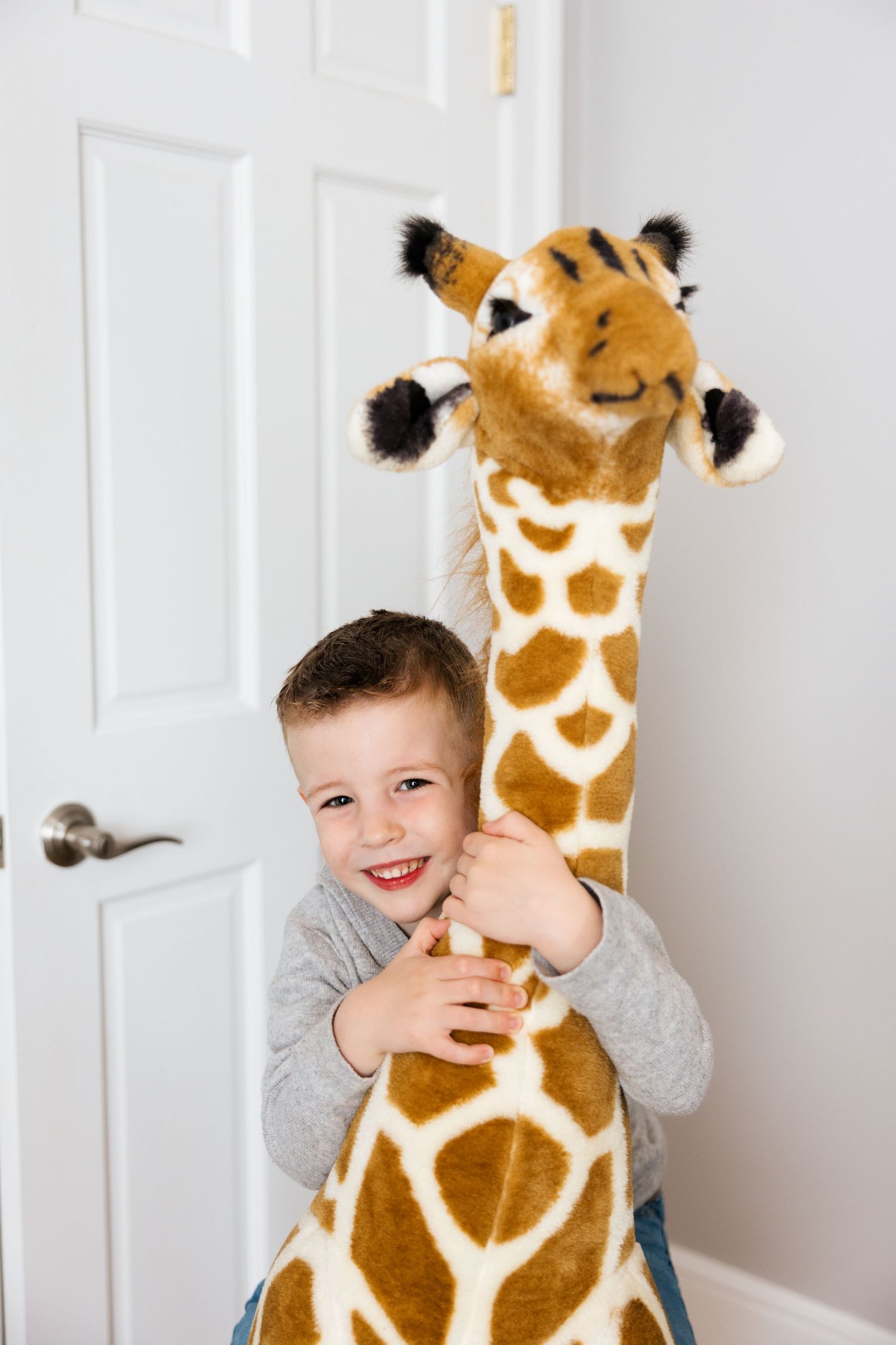 A cheerful young child plays and poses with a large plush giraffe toy against a white wall.