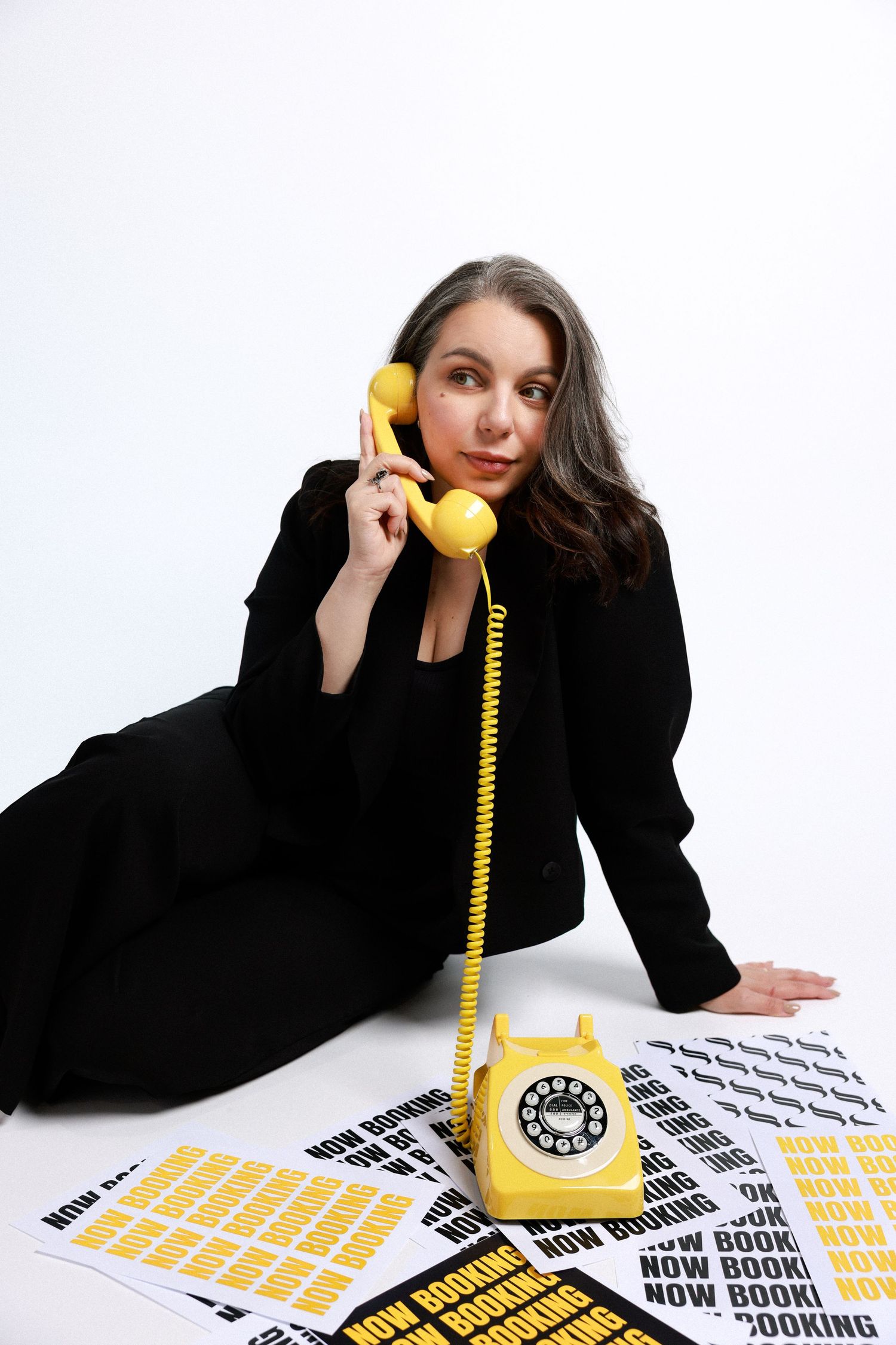 A woman conducts multiple business calls using a bright yellow retro telephone while sitting at a desk with printed papers.