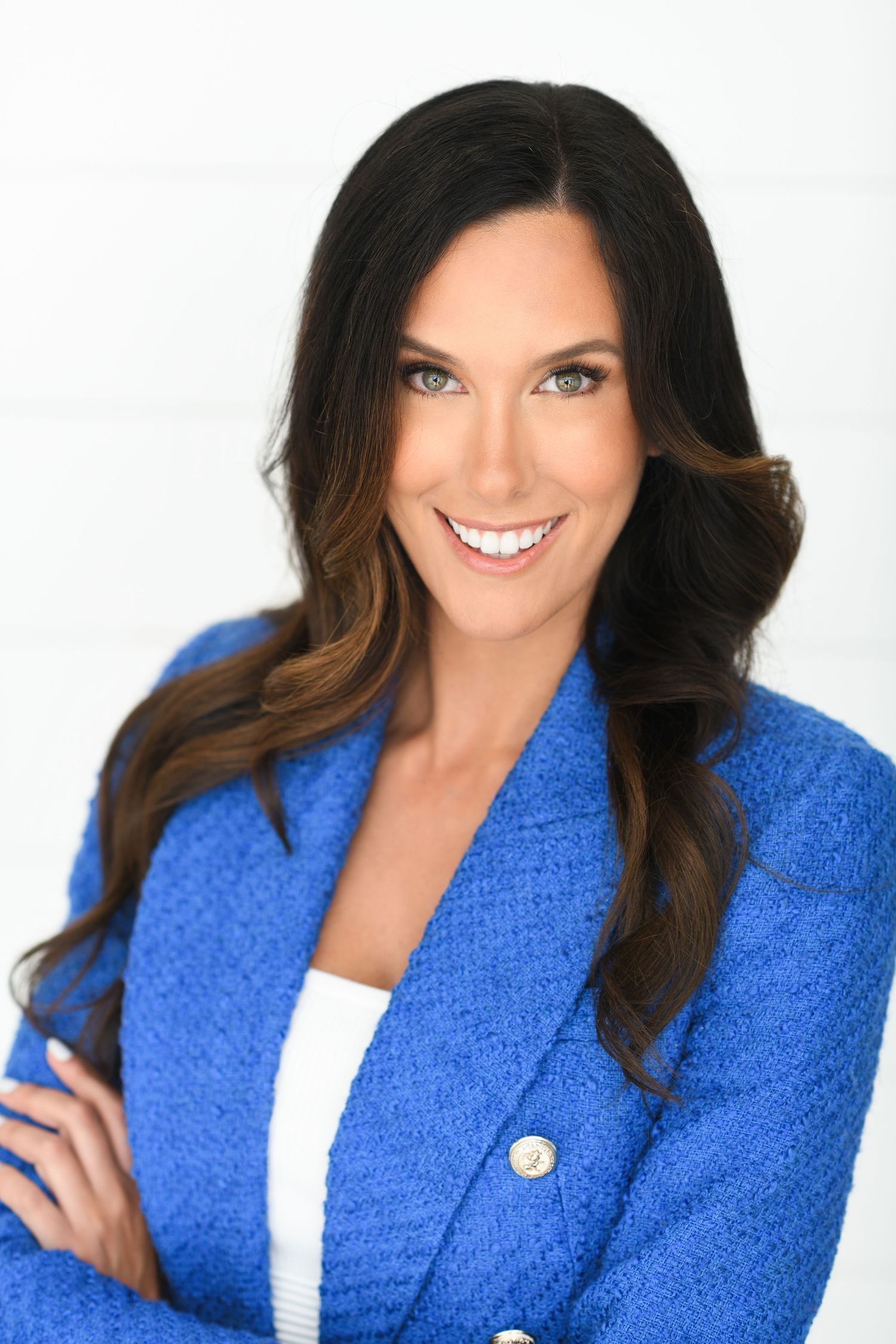 Confident brunette woman in blue blazer with arms crossed smiling against white background.