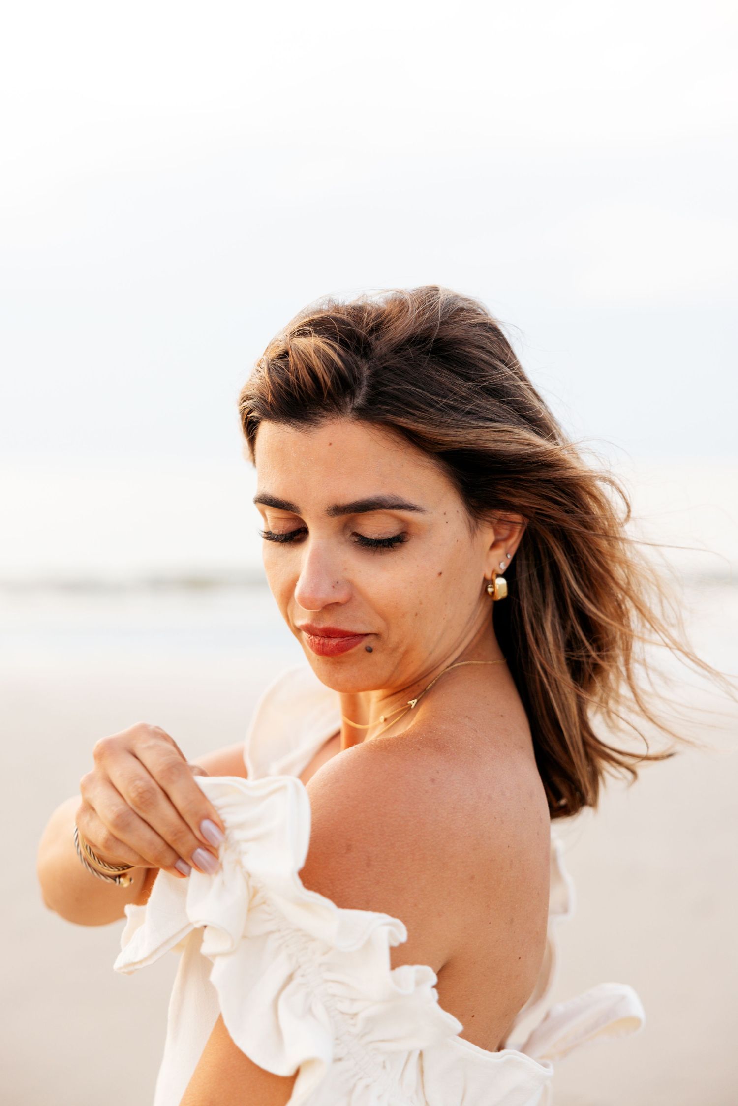 A smiling figure in white wearing a ruffled top poses with windswept hair against a bright outdoor background.