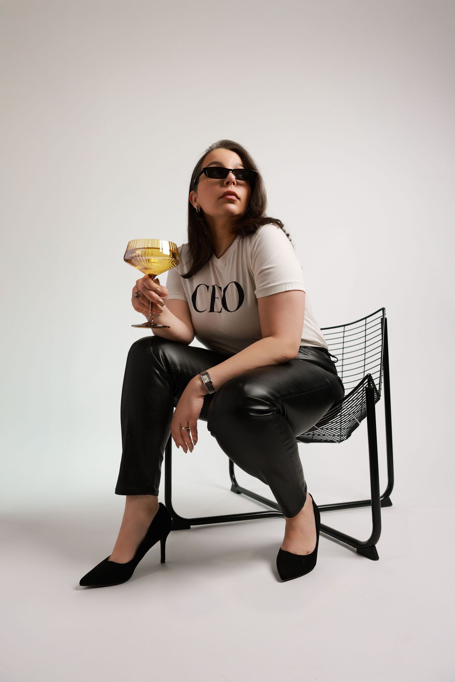 Stylish studio fashion photos show someone in casual attire lounging on a modern black chair with a cocktail glass.
