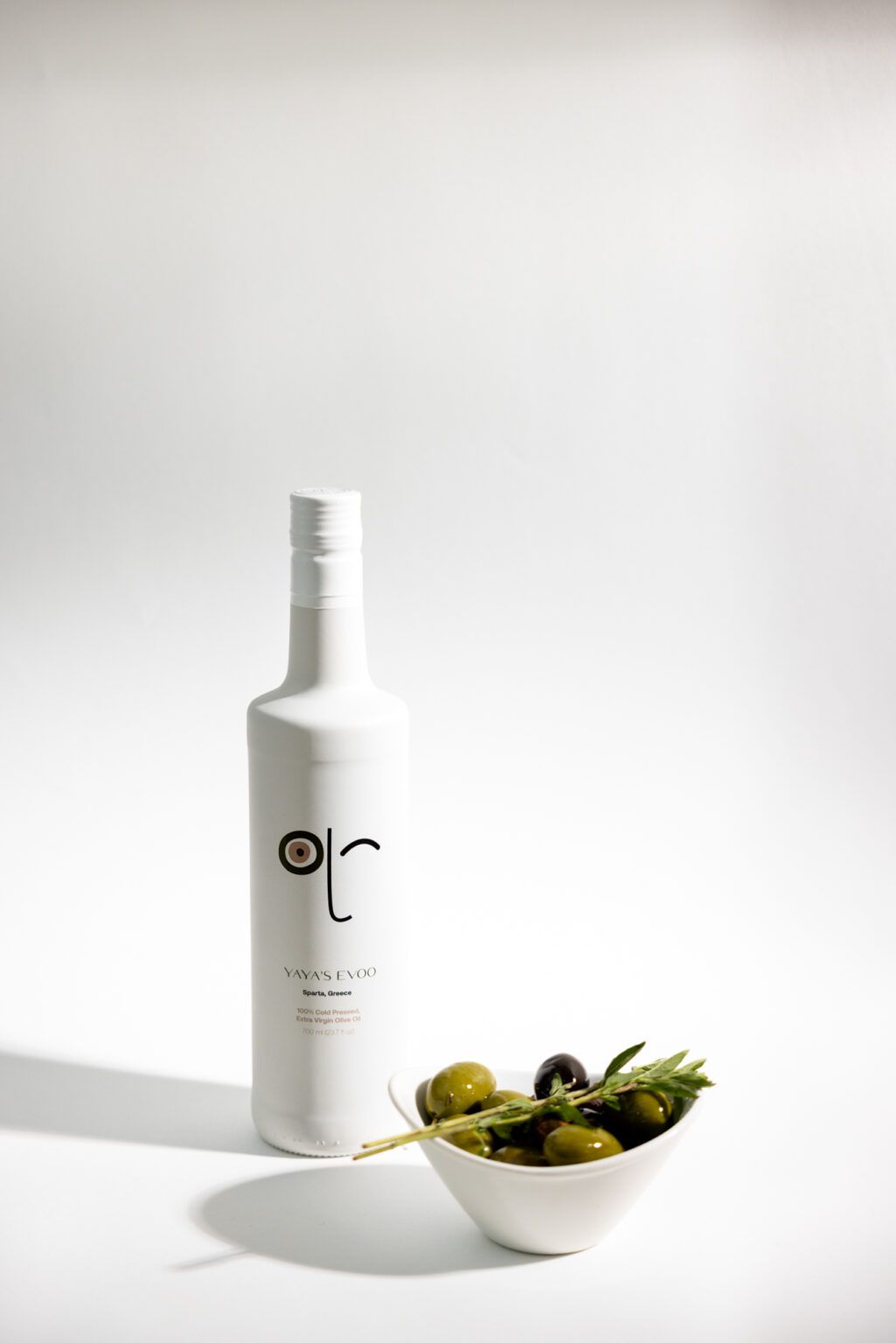 A minimalist white bottle of olive oil next to a small white bowl filled with green olives and rosemary on white surface.