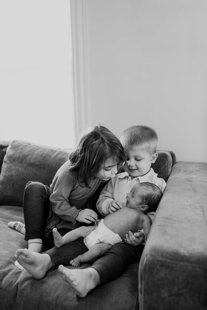 A tender black and white photograph shows siblings cuddling with a newborn baby on a gray sofa.