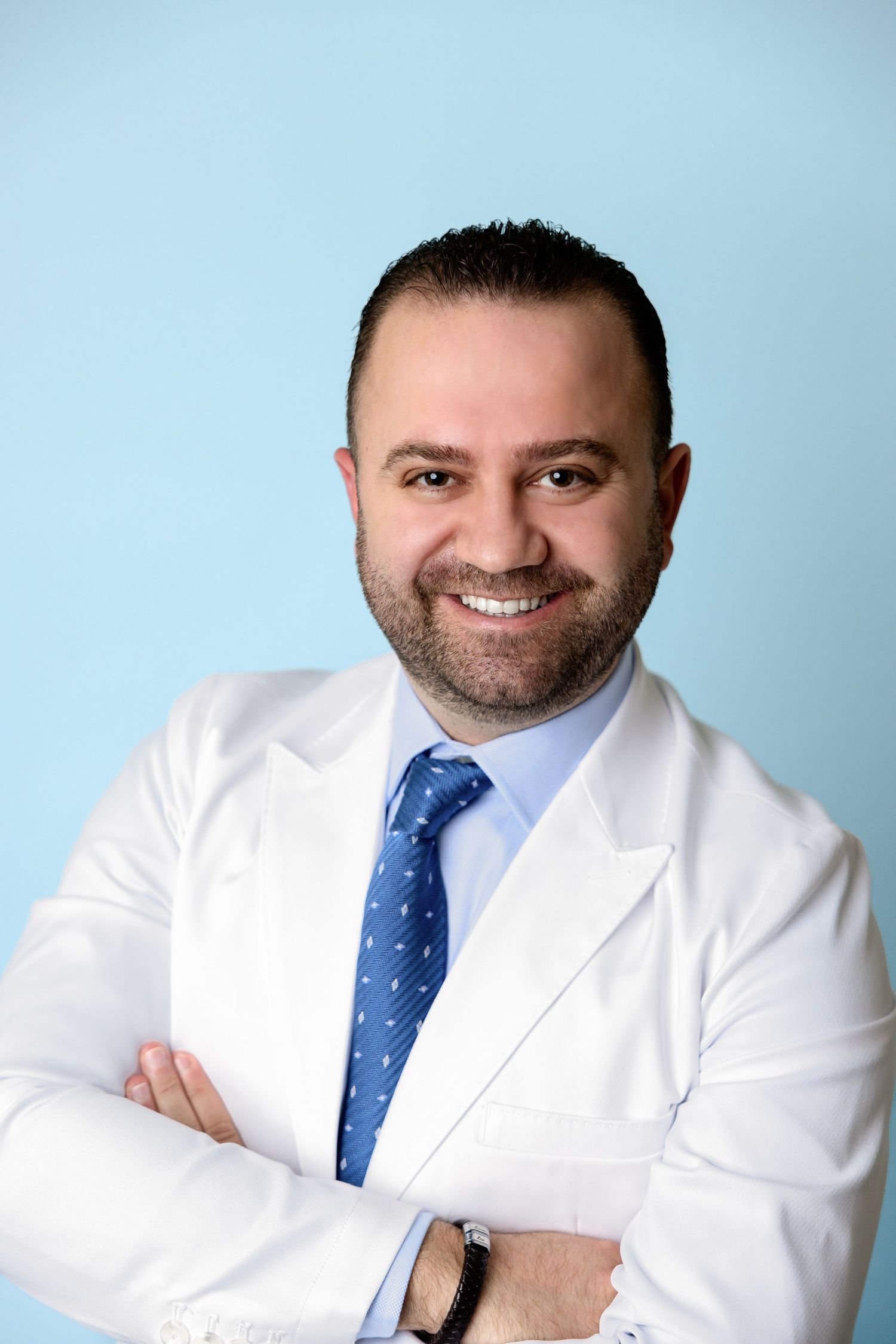 Smiling male doctor in white lab coat and blue tie with arms crossed against light blue background.