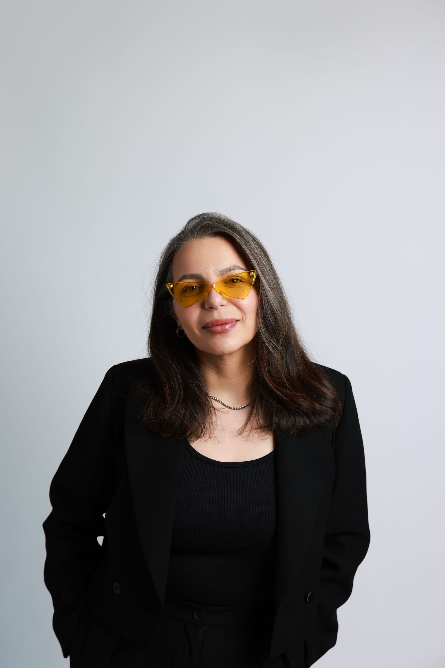 Woman in black blazer and yellow sunglasses smiling against light gray background.