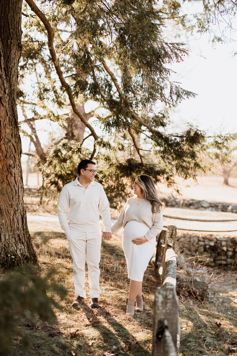 Couple in all white outfits walking together under sunlit trees in a natural setting.
