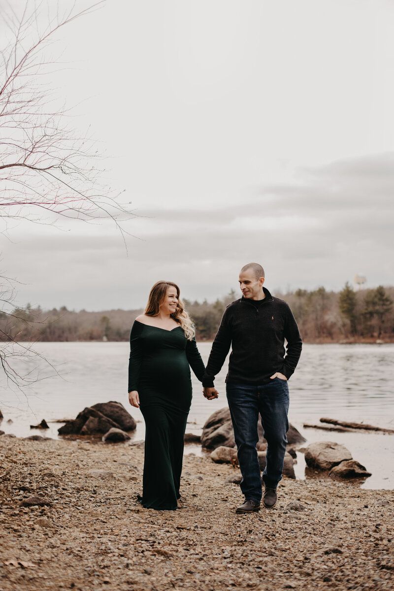 Winter maternity session along a rocky shoreline with bare trees and calm waters.