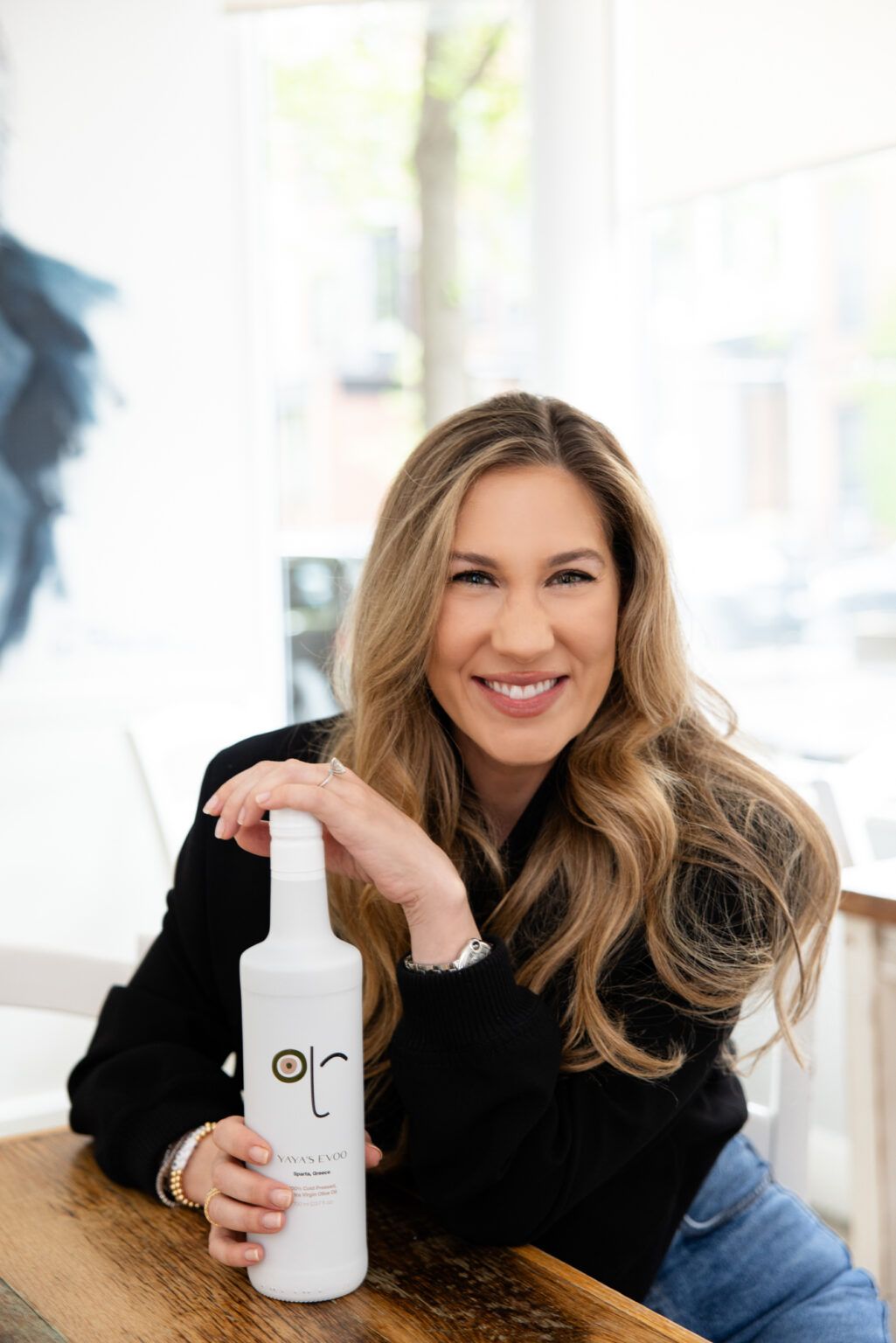 Woman in black sweater holding white coffee mug and smiling while sitting at cafe table.