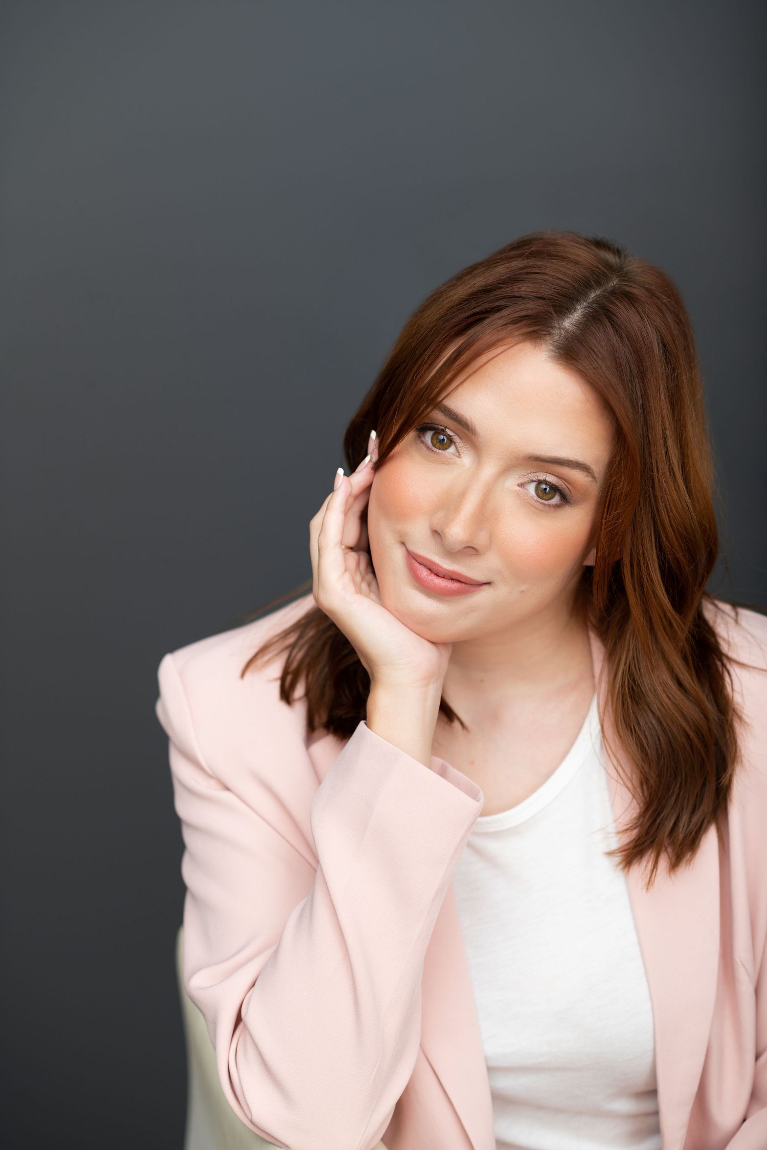 A young woman with auburn hair in a pink blazer poses gracefully with hand near face against gray background.