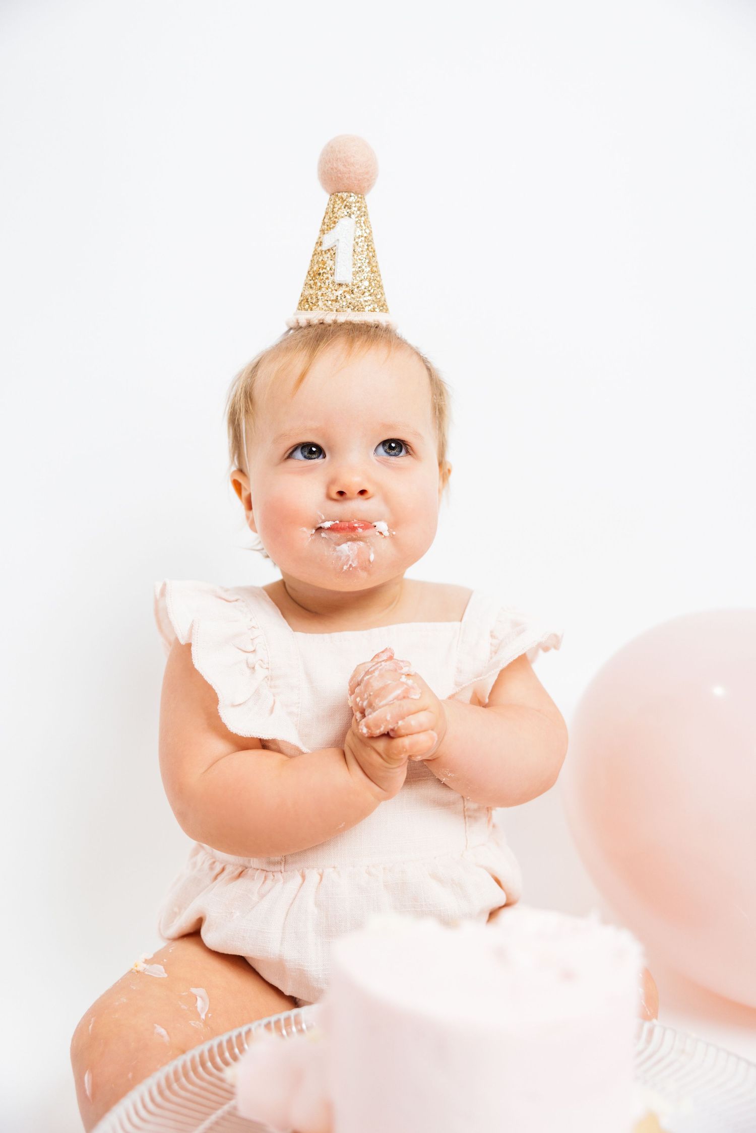 Cute baby girl in white dress and gold birthday hat smiling with cake on face at first birthday party.