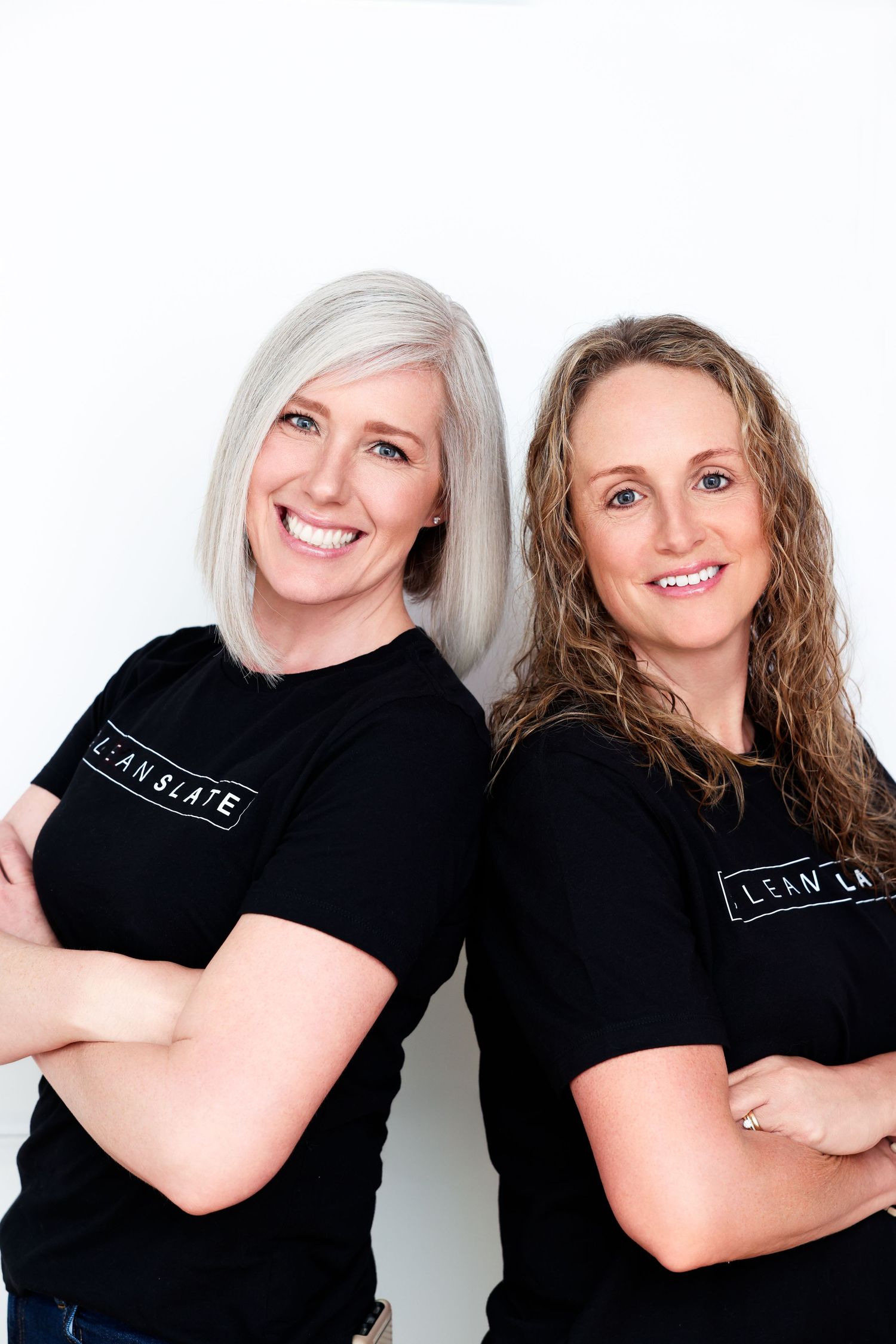 Two smiling women in black Lean Slate t-shirts pose back-to-back with arms crossed against a white background.