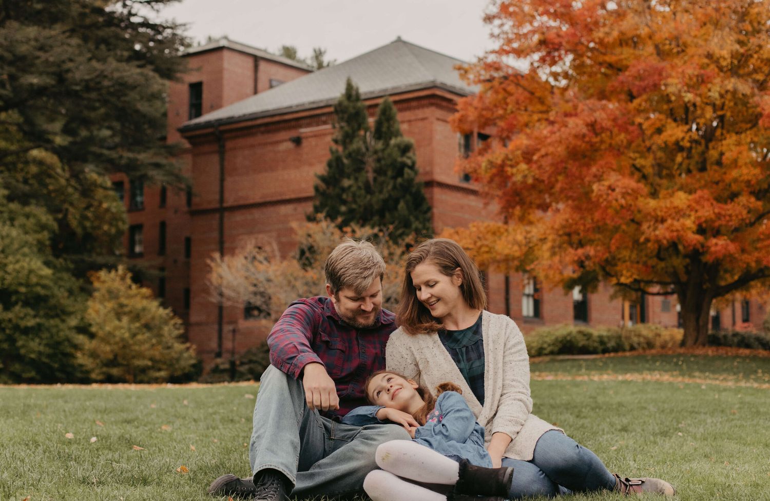 Family relaxing on grass near brick building with fall foliage.